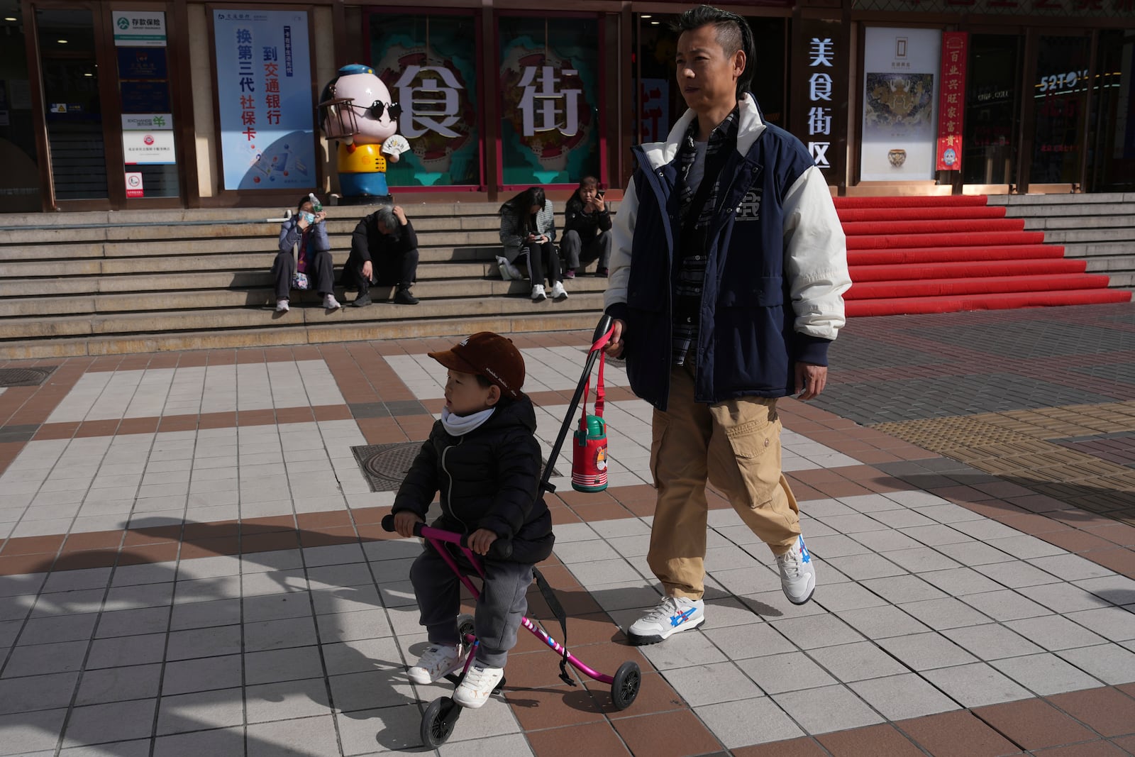 A man pushes a child in a tricycle along the popular Wangfujing shopping street in Beijing, China, Thursday, March 6, 2025. (AP Photo/Ng Han Guan)