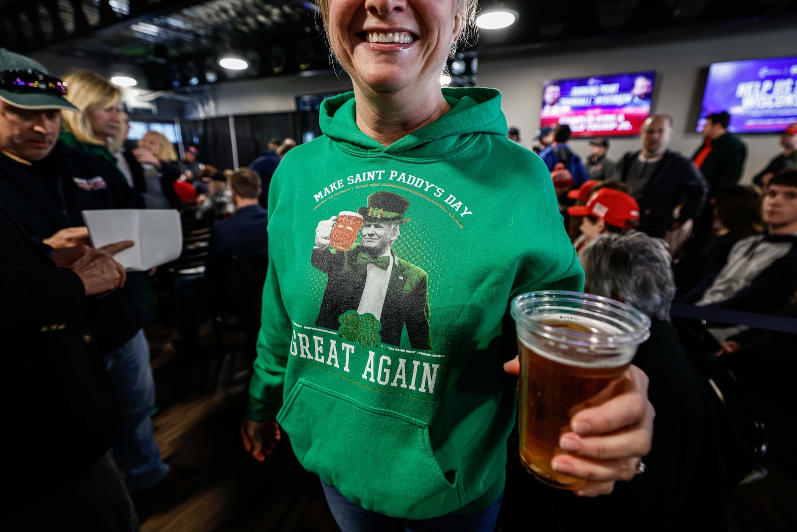 A woman wearing a Donald Trump and Saint Patrick's Day themed hoodie enjoys a beer before a town hall meeting Monday, March 17, 2025, in Oconomowoc, Wis. (AP Photo/Jeffrey Phelps)