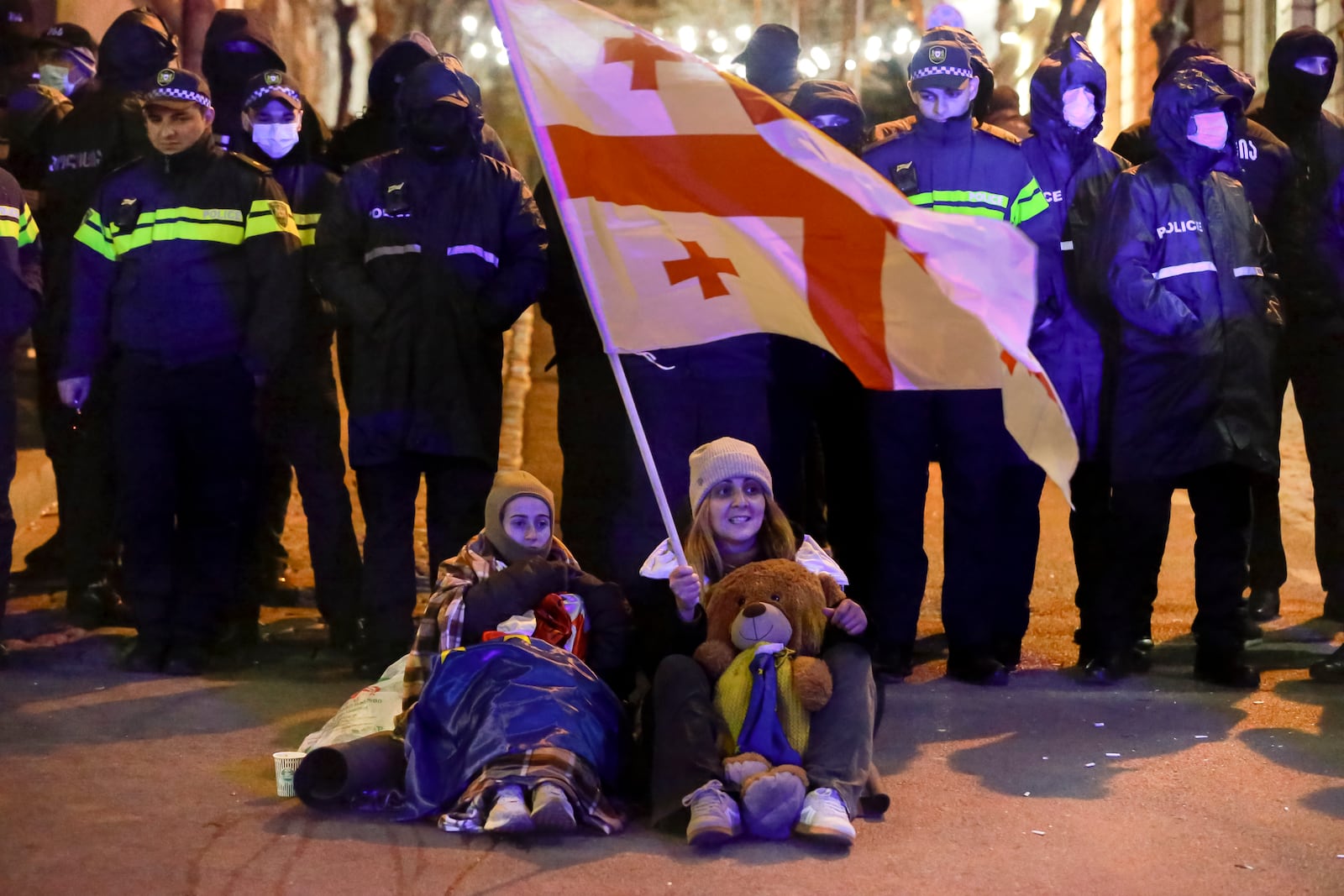 A woman and a girl sit holding a Georgian national flag in front of police blocking the entrance of the Parliament's building during a rally to demand new parliamentary elections in the country, in Tbilisi, Georgia, on Monday, Nov. 25, 2024. (AP Photo/Zurab Tsertsvadze)