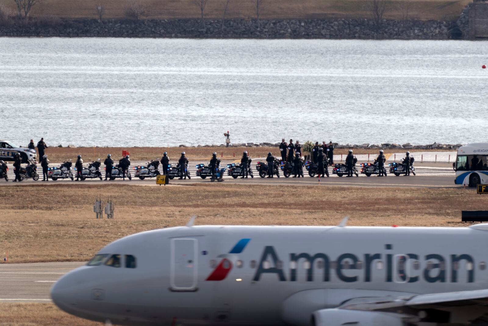 An American Airlines jet passes as police officers escort buses carrying family members of the victims of a mid-air collision between an American Airlines jet and an Army helicopter to runway 33 near the wreckage site in the Potomac River at Ronald Reagan Washington National Airport, Sunday, Feb. 2, 2025, in Arlington, Va. (AP Photo/Jose Luis Magana)