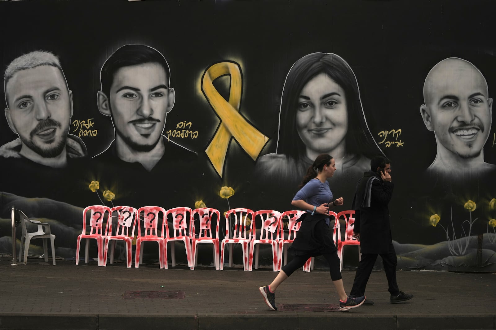 People walk and jog past an installation and a mural of portraits of hostages held by Hamas in the Gaza Strip, in Jerusalem, Friday, Jan. 24, 2025. (AP Photo/Mahmoud Illean)