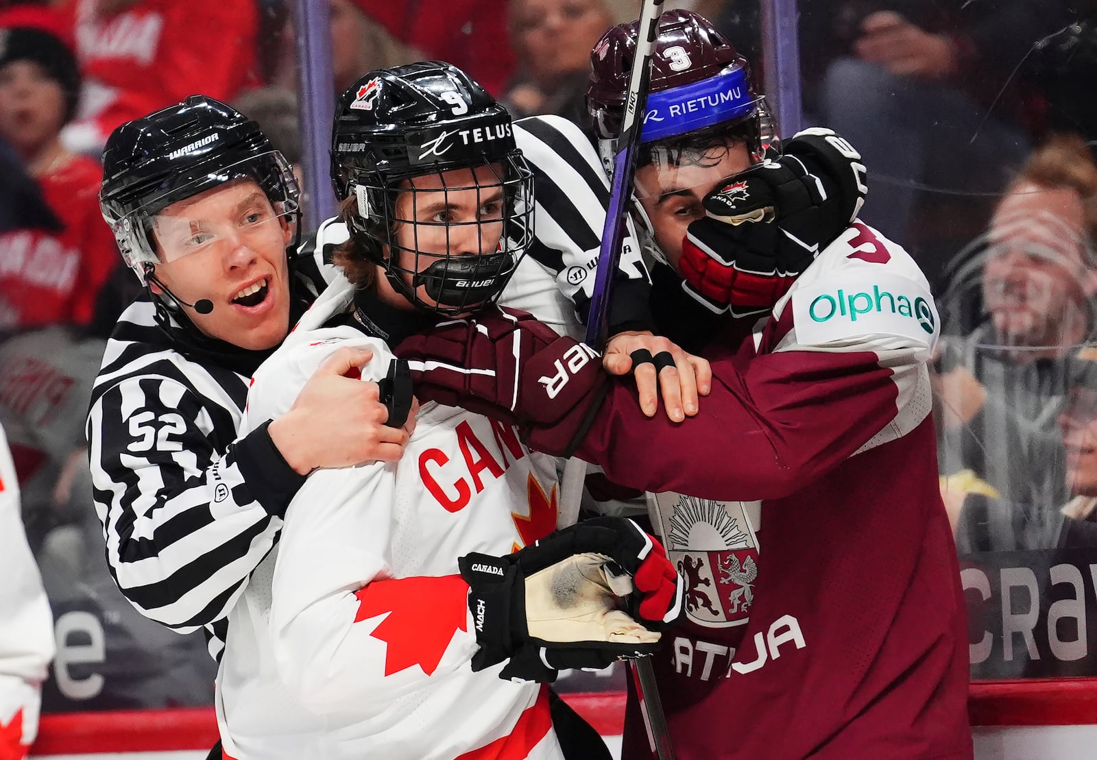 Canada's Gavin McKenna (9) and Latvia's Viktors Kurbaka (3) grab on to each other after the whistle as linesperson Nick Briganti tries to separate them during second period IIHF World Junior Hockey Championship preliminary round actio in Ottawa, Ontario, Friday, Dec. 27, 2024. (Sean Kilpatrick/The Canadian Press via AP)