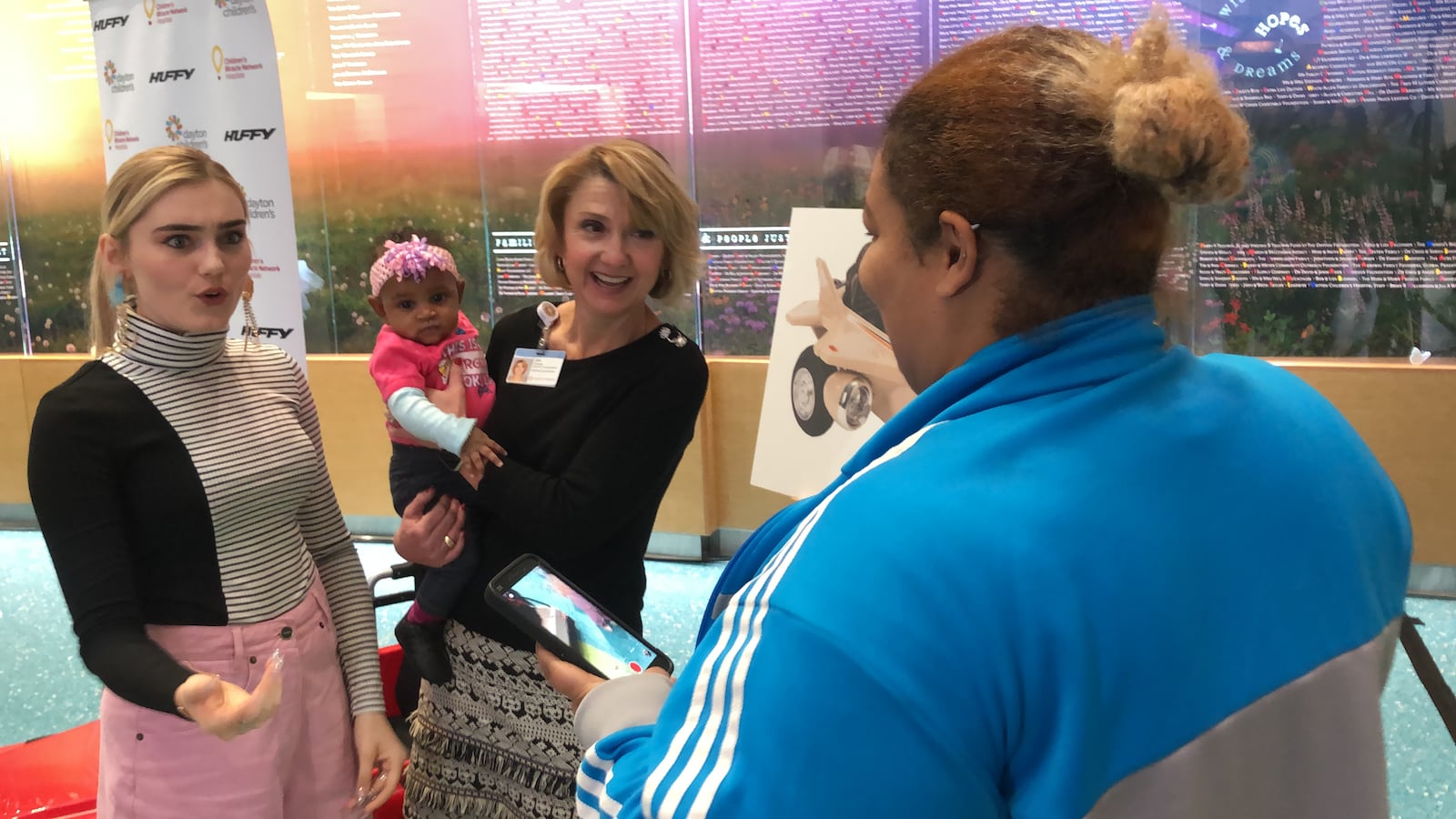 Meg Donnelly, star of  ABC’s  “American Housewife” and the popular Disney Channel movie “Zombies” smiles at 5-month-old Rai’Mia Bass being held by Julie Cannon of Dayton Children’s Hospital.  Rai’Mia had just finished a chemotherapy treatment. The baby’s mother, Brittany Bass, records a message for her 6-year-old daughter, a Donnelly fan. Donnelly visited Dayton Children’s as part of the announcement of Huffy’s donation to  Dayton Children’s and other hospitals in the Children’s Miracle Network. Photo by Amelia Robinson