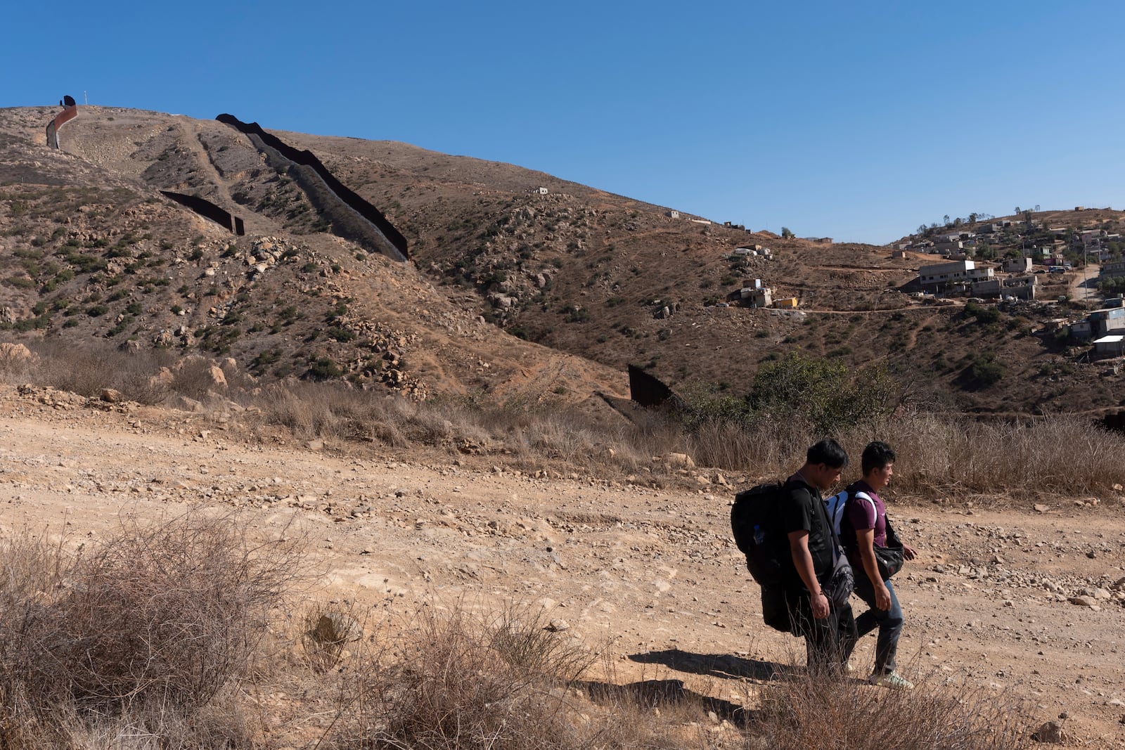 Two men walk down a road towards Border Patrol agents after crossing the border illegally through a gap in two walls separating Mexico from the United States, behind, before turning themselves in, Thursday, Jan. 23, 2025, in San Diego. (AP Photo/Gregory Bull)