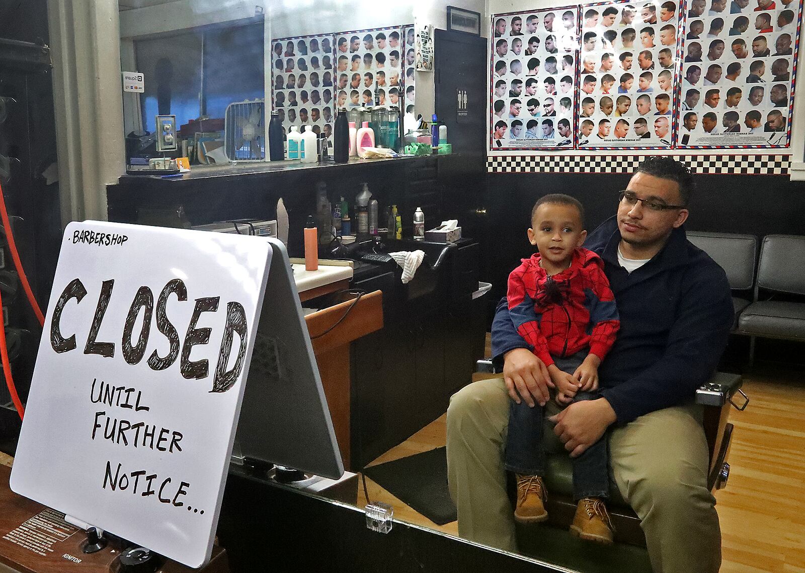 Alex Robinson, owner of Robinson Corner Cuts, and his son, Jarred, sit in his empty barbershop on March 18 after the governor ordered all barbershops and nail salons to close Wednesday due to the coronavirus. BILL LACKEY/STAFF