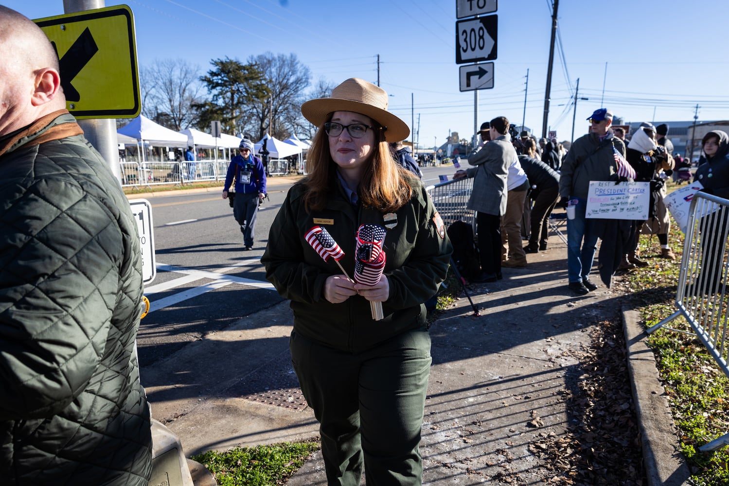 A park service officer hands out American flags to people gathered near the main street of Plains, Ga., on Jan. 4, 225, before a funeral procession for President Jimmy Carter. (Dustin Chambers/The New York Times)