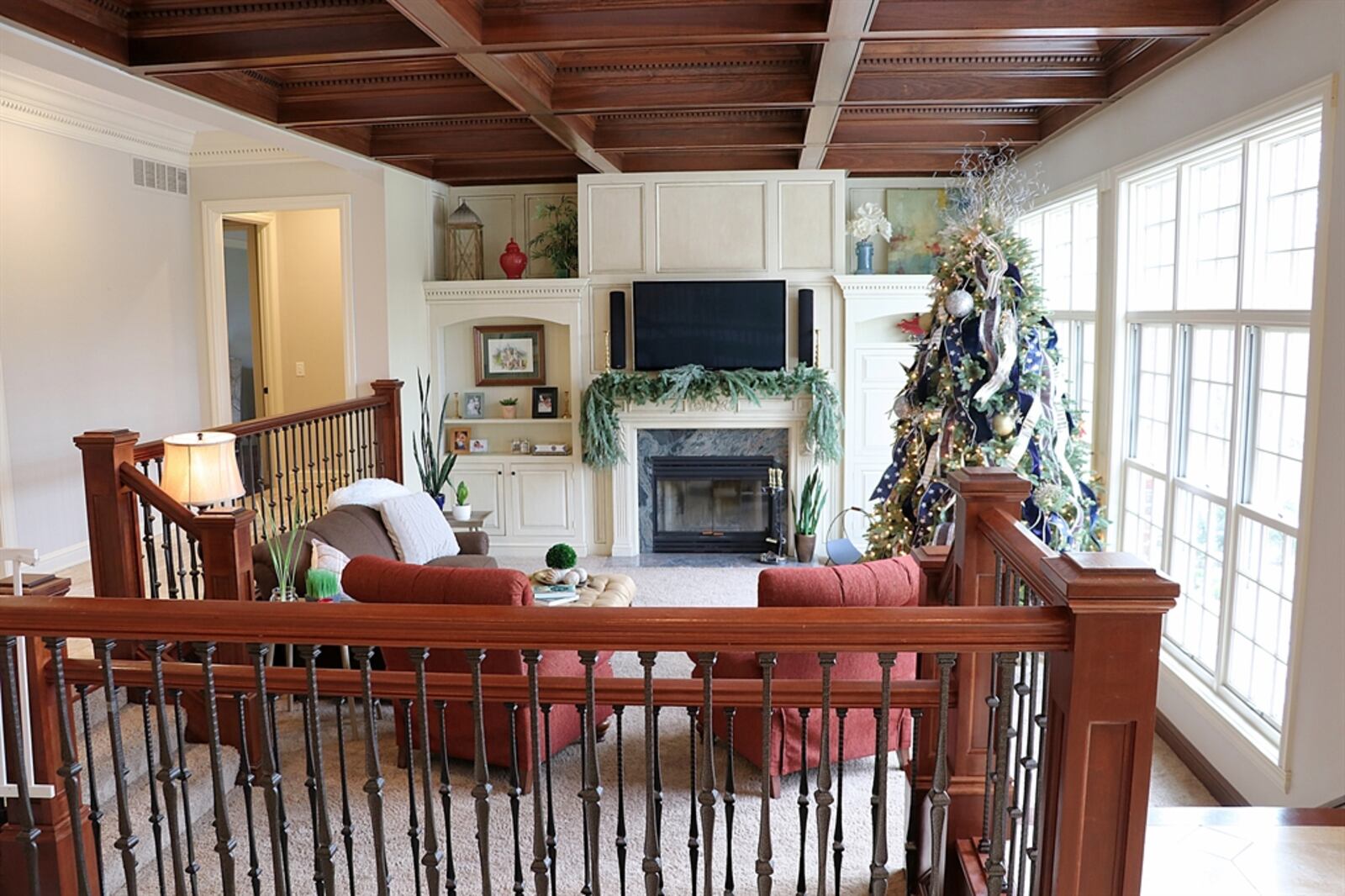 Coffered ceiling and wrought-iron railing accent the sunken great room, which has a wall of windows that looks out over the pool area. Along another wall is off-white cabinetry that flanks a gas fireplace.