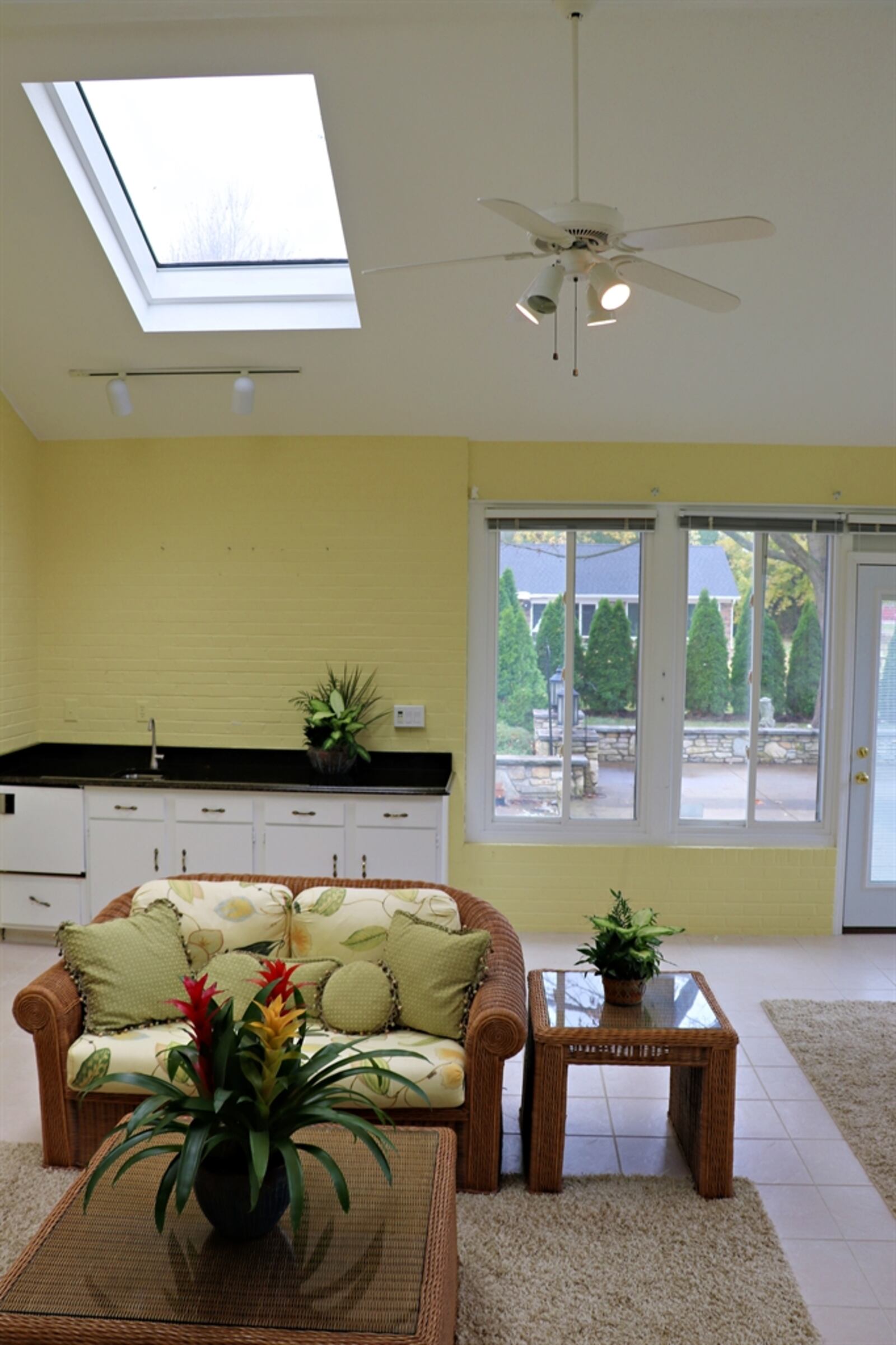 A cathedral ceiling with skylights peaks above the four-season room, which has a wall of windows and access to the courtyard patio. Windows wrap around one corner and end at a long counter bar with white cabinets beneath. The counter bar has a sink and an appliance nook.