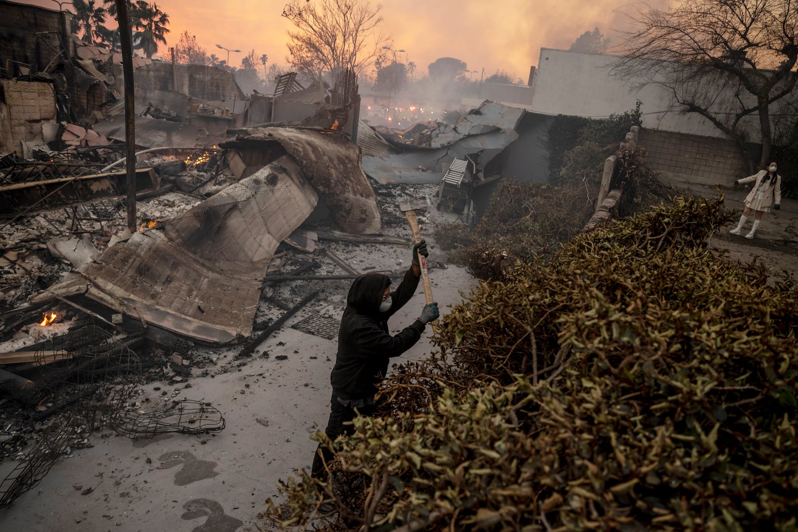 A person cuts down bushes as structures are seen on fire during the Eaton fire in Altadena, Calif., Wednesday, Jan. 8, 2025. (Stephen Lam/San Francisco Chronicle via AP)