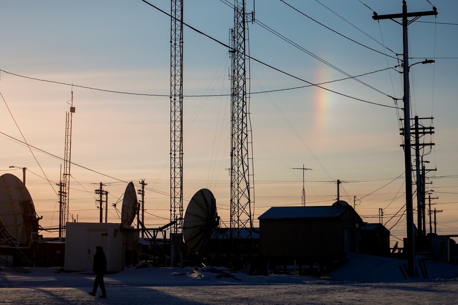 A sundog forms a vertical rainbow as a villager walks by transmission dishes in Kaktovik, Alaska, Tuesday, Oct. 15, 2024. (AP Photo/Lindsey Wasson)