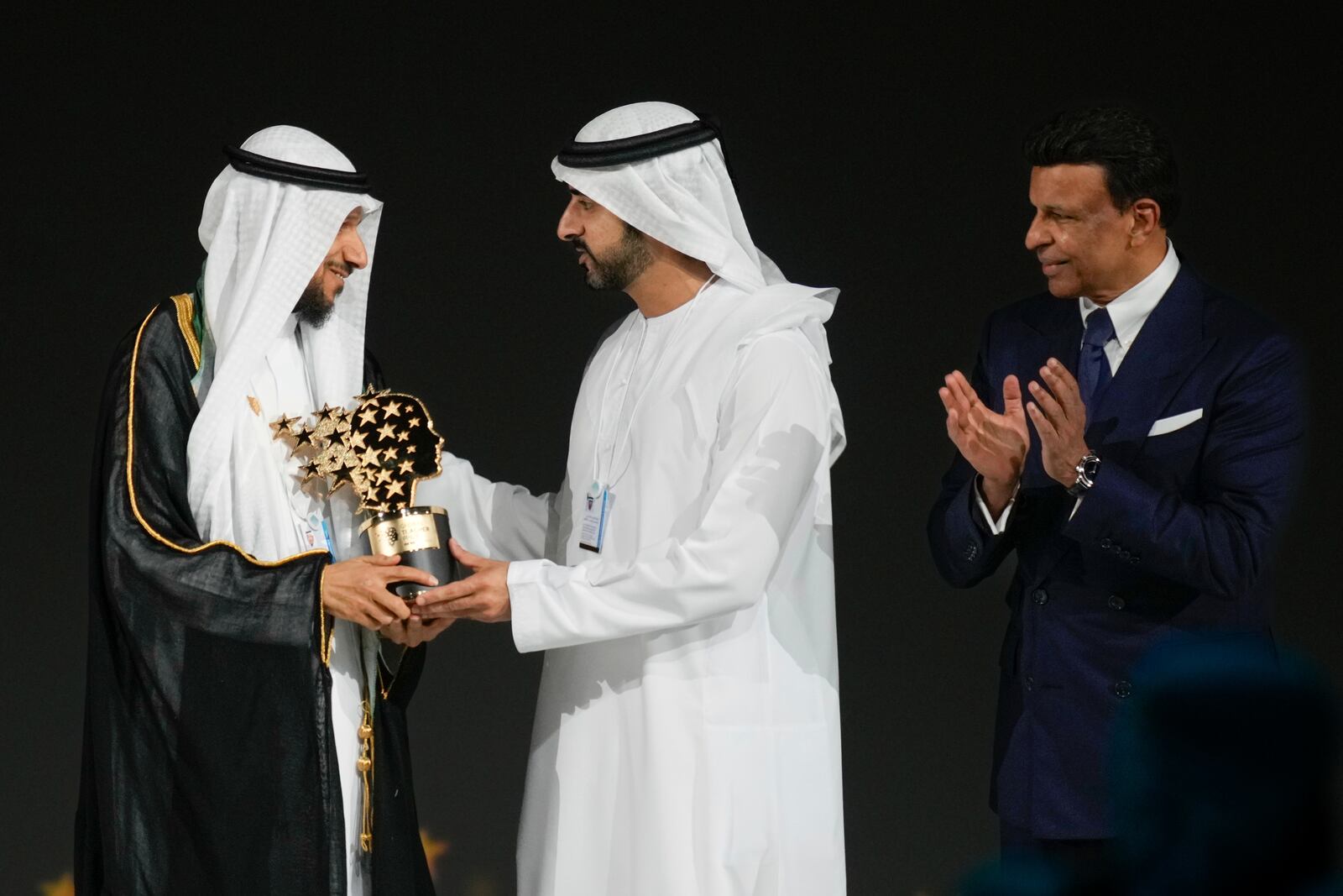 Saudi Arabian teacher Mansour bin Abdullah Al-Mansour, left, receives the Global Teacher Prize trophy from Dubai Crown Prince Sheikh Hamdan bin Mohammed Al Maktoum as Varkey foundation founder Sunny Varkey, right, applauds at a ceremony awarding the Global Teacher Prize in Dubai, United Arab Emirates, Thursday, Feb. 13, 2025. (AP Photo/Altaf Qadri)