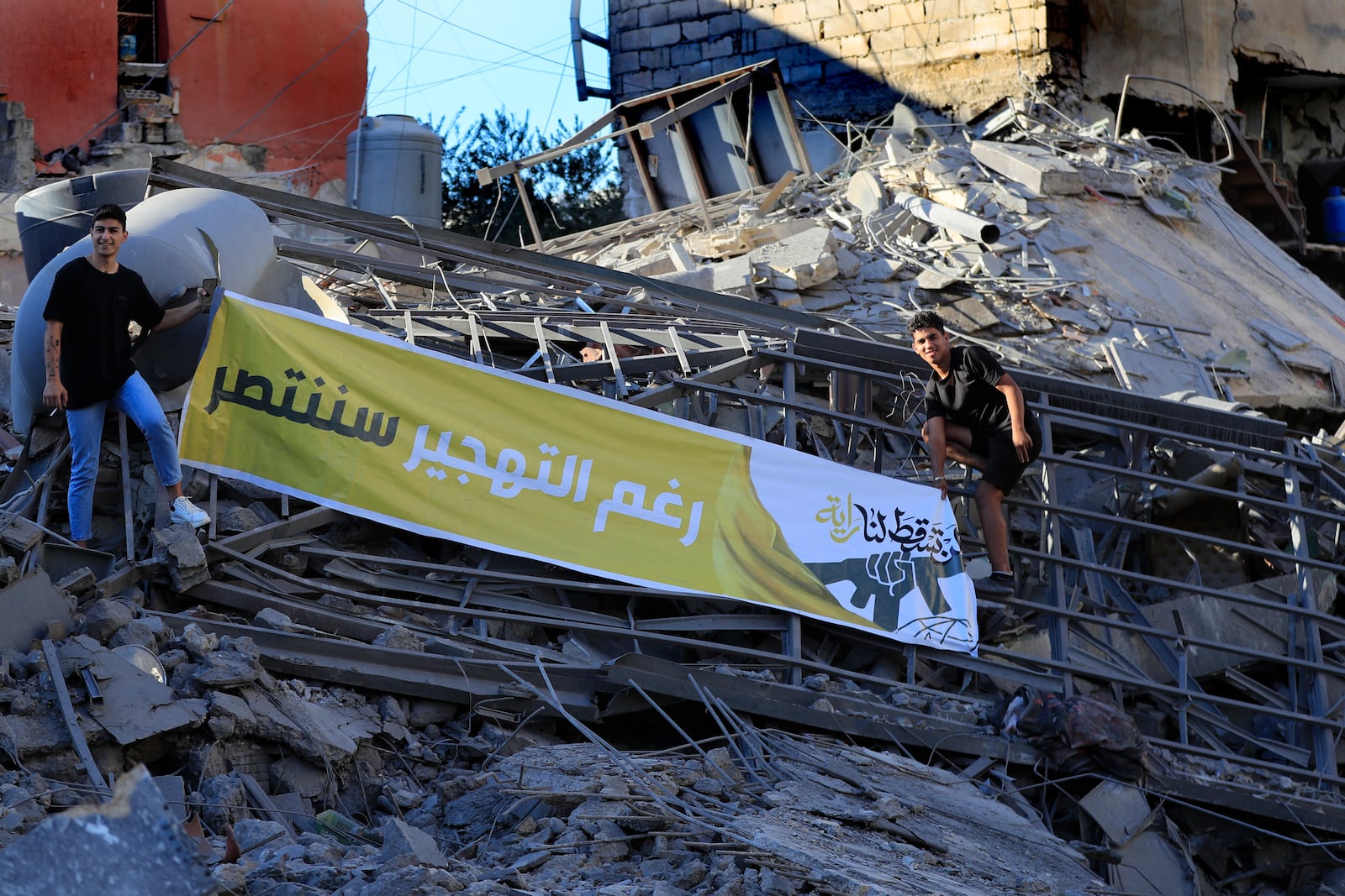 Hezbollah supporters stand on the rubble of a destroyed building hit by Israeli airstrikes, as they hold an Arabic banner that reads: "Despite the displacement we will be victorious", in Tyre, south Lebanon, Wednesday, Oct. 23, 2024. (AP Photo/Mohammed Zaatari)