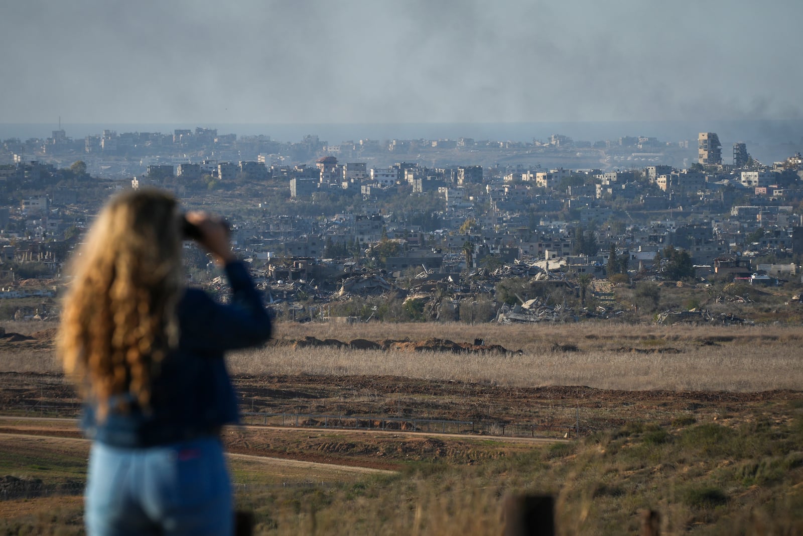 A woman uses binoculars to watch smoke rising following an explosion in the Gaza Strip as seen from southern Israel, Wednesday, Dec.18, 2024. (AP Photo/Ohad Zwigenberg)