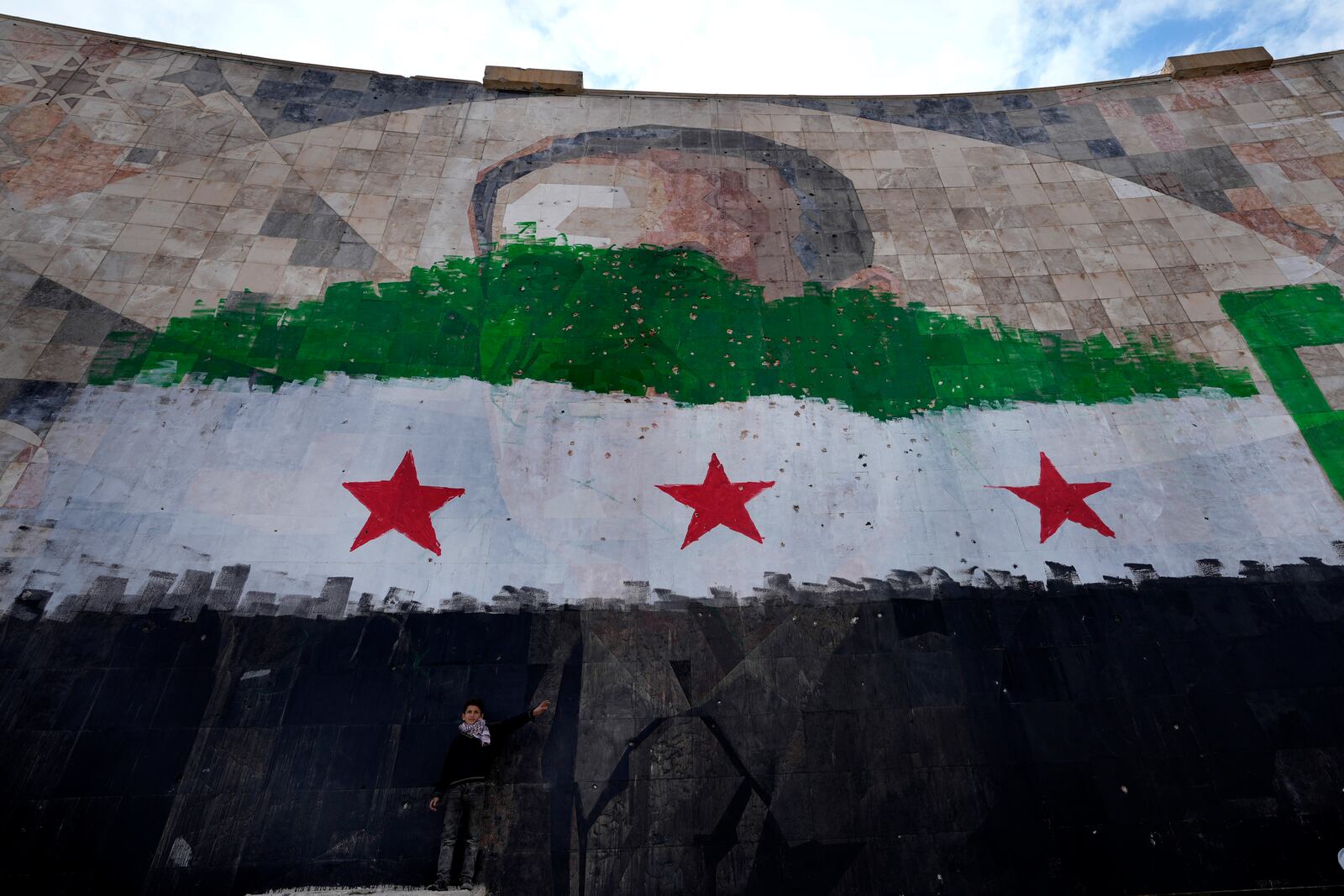 A Syrian boy stands under a giant portrait of the late Syrian President Hafez Assad painted over with the colors of the "revolutionary" flag, in Damascus, Syria, Thursday, Dec. 26, 2024. (AP Photo/Hussein Malla)