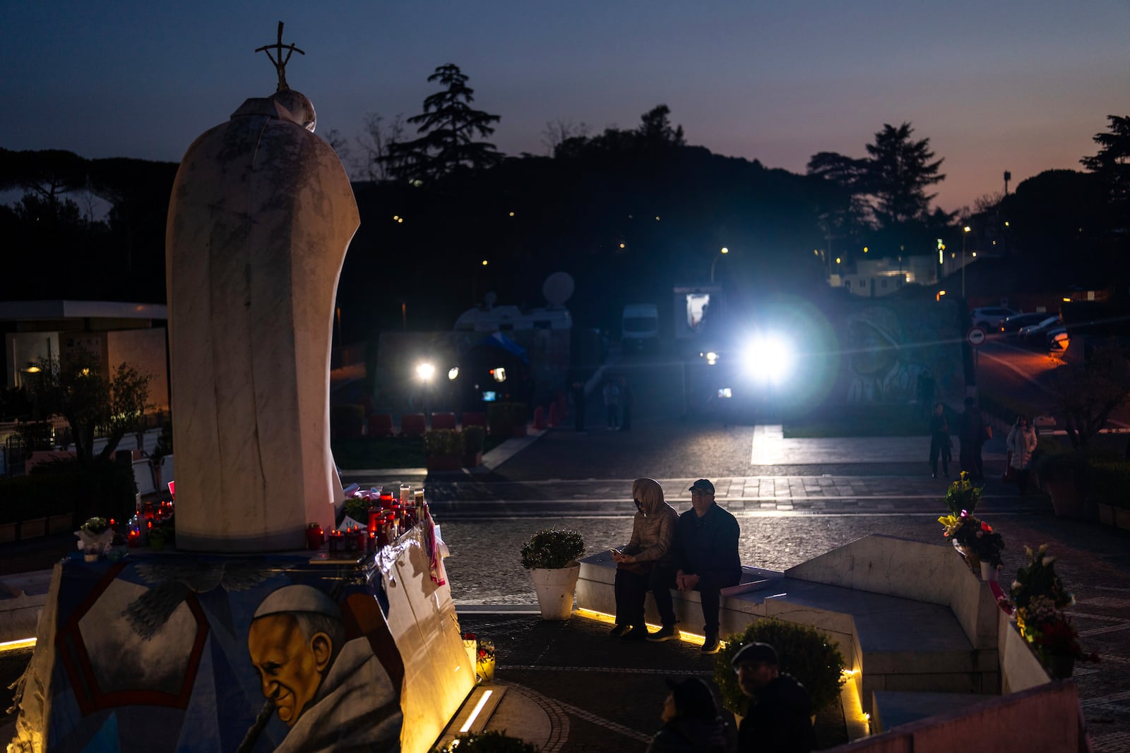 People sit next to the statue of Pope John Paul II placed outside the Agostino Gemelli hospital in Rome, Thursday, March 6, 2025. (AP Photo/Francisco Seco)