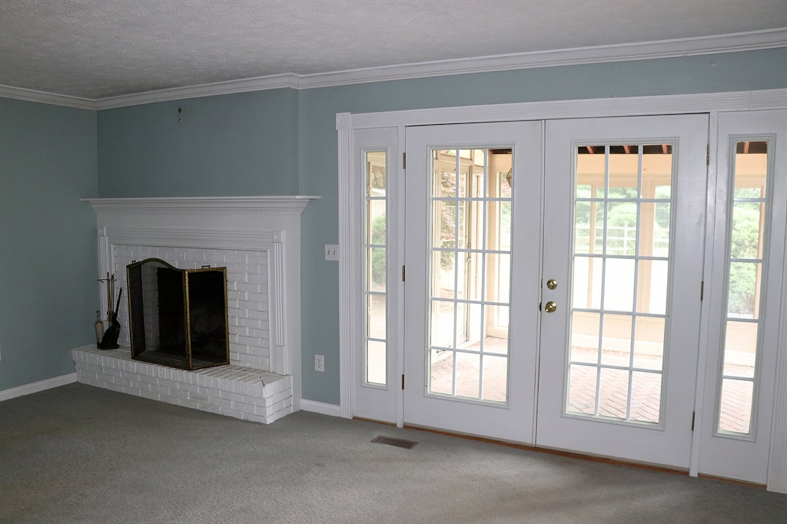 A spacious family room has a wood-burning fireplace tucked near a corner. The fireplace has a raised brick hearth and dentil wood mantel. Next to the fireplace, French doors allow access to an enclosed rear porch.