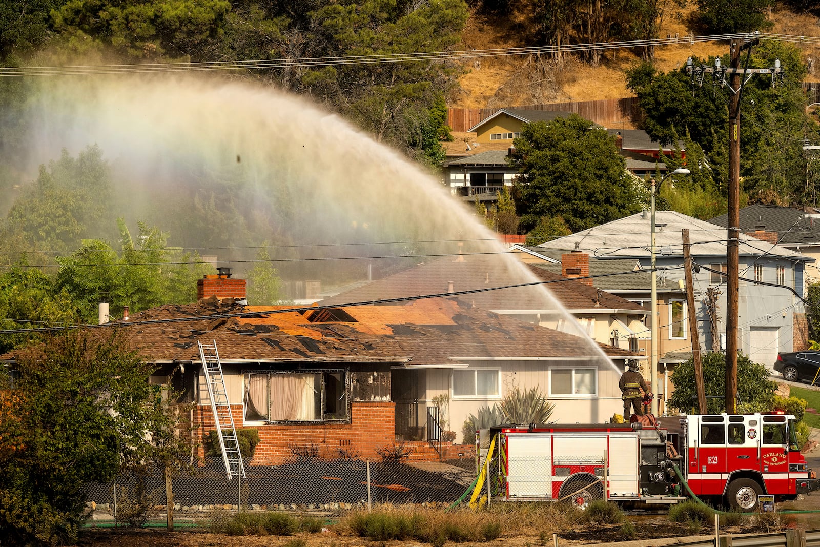 A firefighter sprays water on a scorched home as a grass fire burns above Interstate 580 in Oakland, Calif., Friday, Oct. 18, 2024. (AP Photo/Noah Berger)