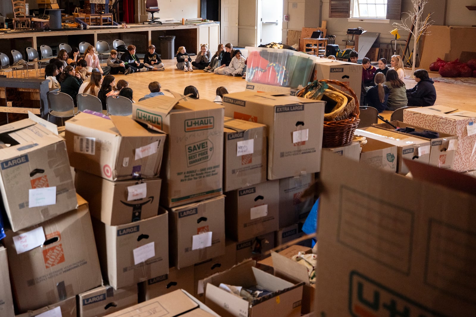 Containers filled with donations for the people impacted by the wildfires are stacked in the back as students with the Theatre Palisades' youth theatre program participate in a master class with American actress and singer Kerry Butler after their theater was destroyed by the Palisades Fire, at the Saint Monica Preparatory's auditorium in Santa Monica, Calif., Sunday, Jan. 26, 2025.(AP Photo/Etienne Laurent)