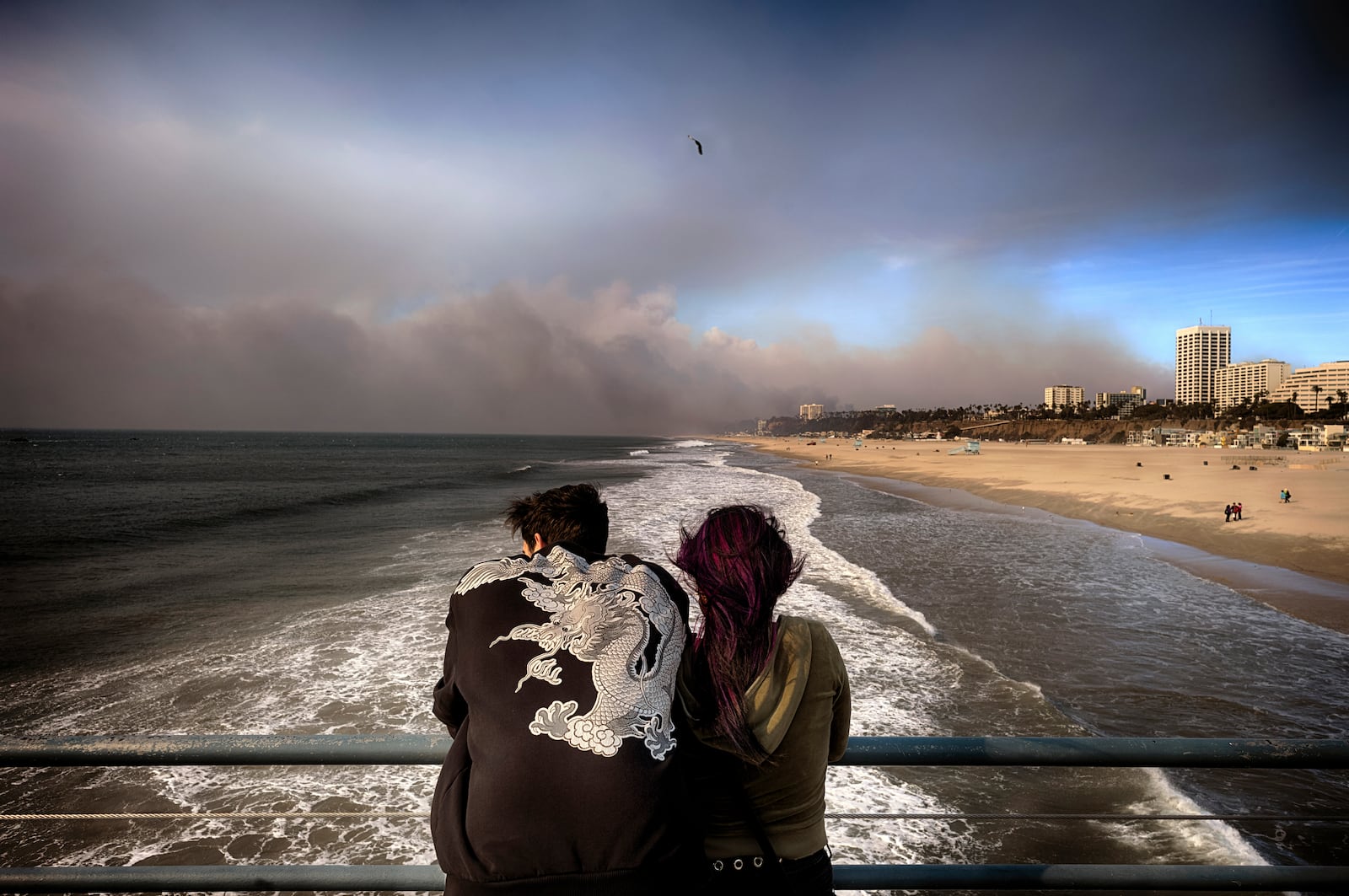 Visitors to the Santa Monica pier look out at smoke from a wildfire in the Pacific Palisades blows over the beach in Santa Monica, Calif., on Wednesday, Jan. 8, 2025. (AP Photo/Richard Vogel)