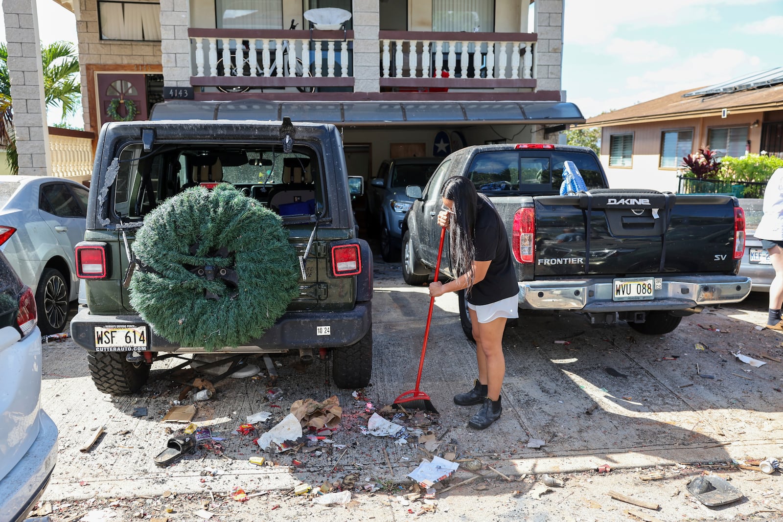 A woman sweeps debris from a driveway across the street from the home where a New Year's Eve fireworks explosion killed and injured people, Wednesday, Jan. 1, 2025, in Honolulu. (AP Photo/Marco Garcia)