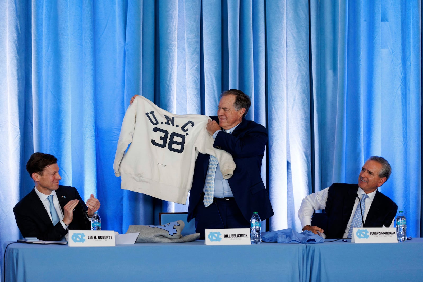 New North Carolina coach Bill Belichick, center, holds up a sweatshirt that belonged to his father when he was on the coaching staff at UNC in the 1950s as Chancellor Lee Roberts, left, and athletic director Bubba Cunningham, right, look on. during an NCAA college football press conference in Chapel Hill, N.C., Thursday, Dec. 12, 2024. (AP Photo/Ben McKeown)