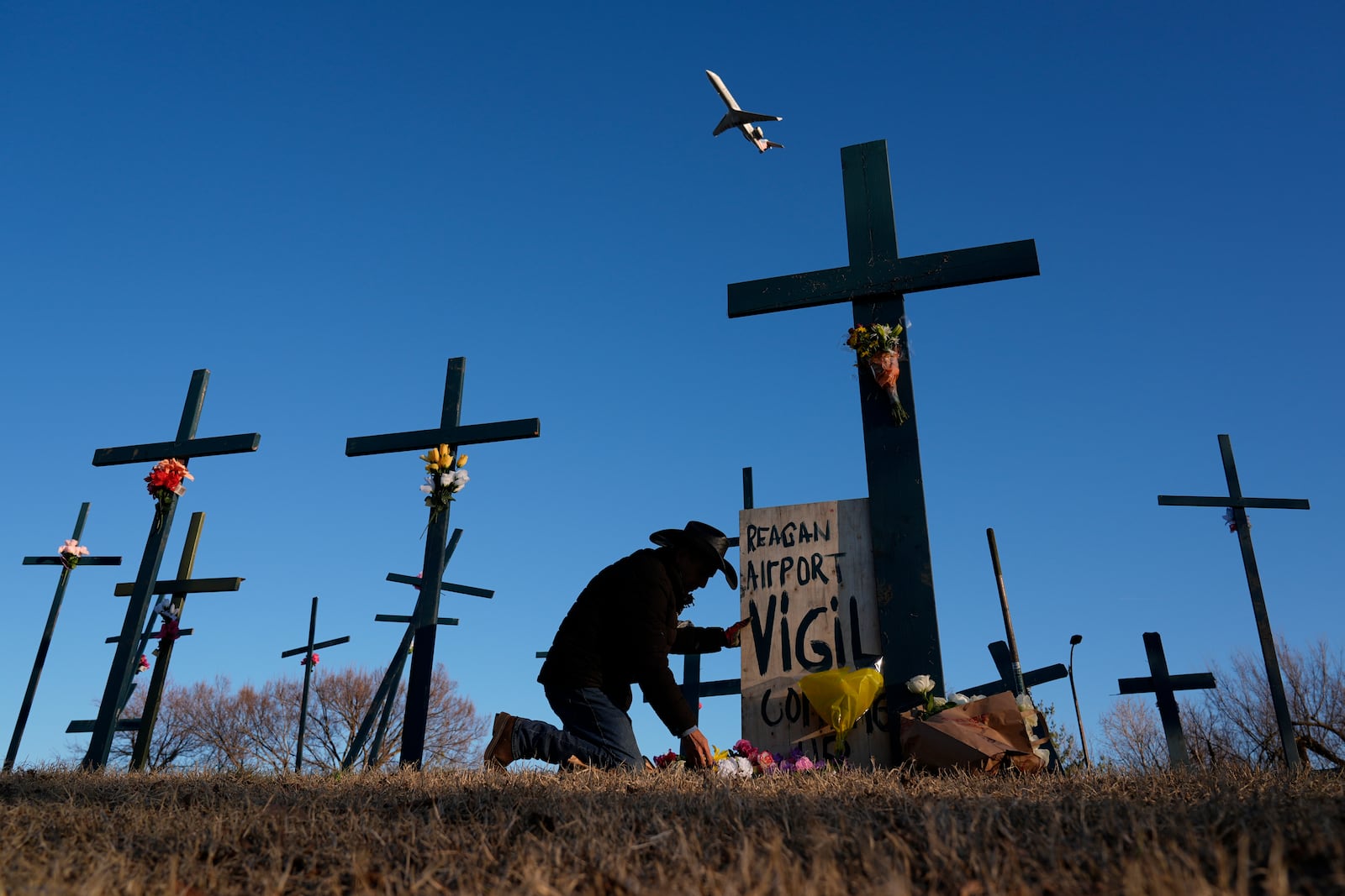 A plane takes off from Ronald Reagan Washington National Airport as Roberto Marquez of Dallas places flowers at a memorial of crosses he erected for the 67 victims of a midair collision between an Army helicopter and an American Airlines jet, Saturday, Feb. 1, 2025, in Arlington, Va. (AP Photo/Carolyn Kaster)