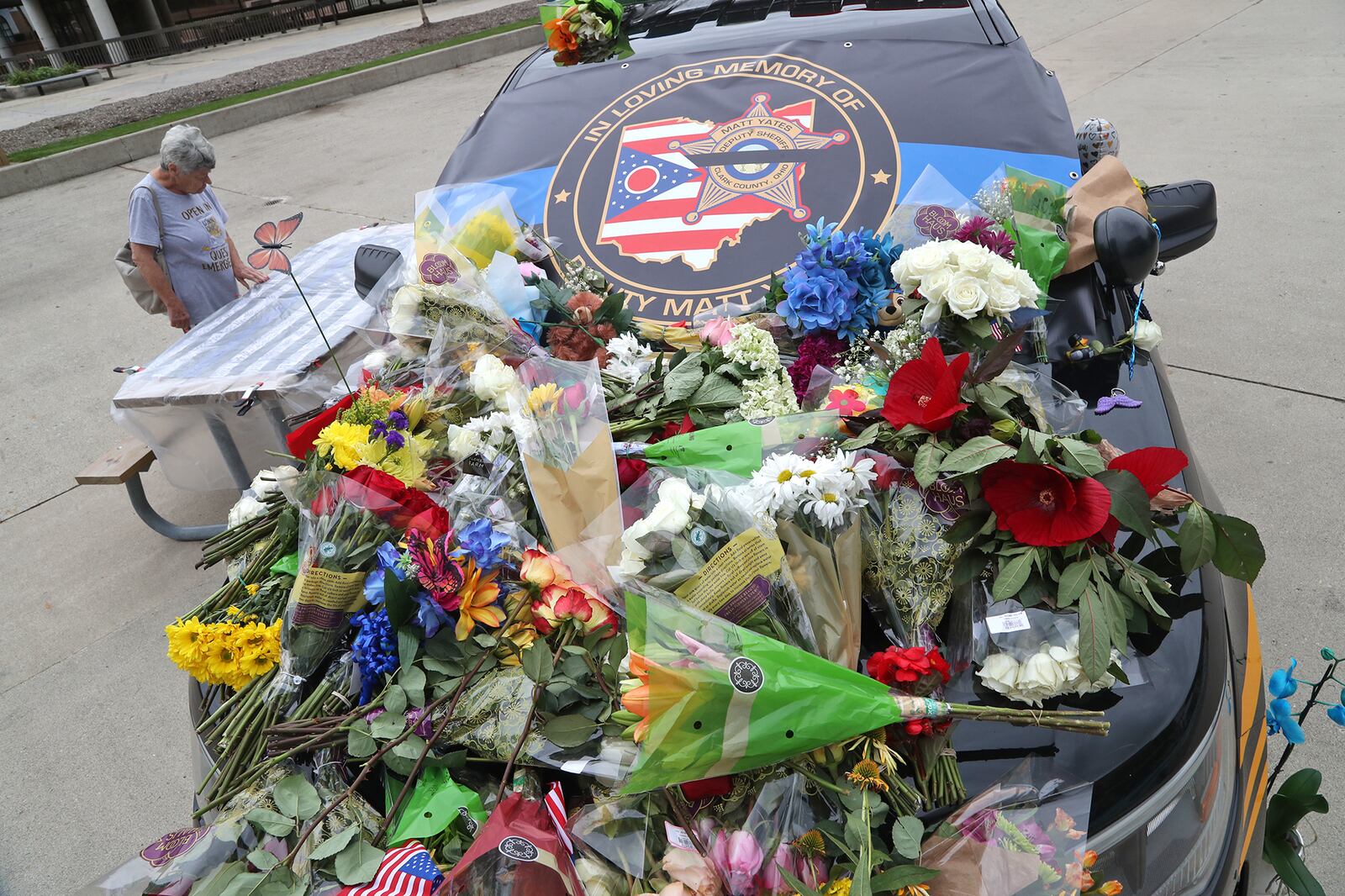 Loretta Martin looks over the messages written on a flag displayed next to a Clark County Sheriff's vehicle covered in flowers on the Springfield City Hall Plaza Tuesday, July 26, 2022. The vehicle is a memorial for Deputy Matthew Yates, who was killed Sunday on a call at Harmony Estates. BILL LACKEY/STAFF