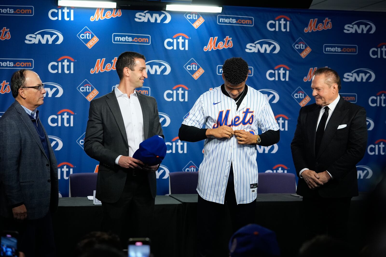 New York Mets' Juan Soto, second from right, puts on a jersey as sports agent Scott Boras, right, Mets owner Steven Cohen, left, and president of baseball operations David Stearns, second from left, watch during a baseball news conference, Thursday, Dec. 12, 2024, in New York. (AP Photo/Frank Franklin II)