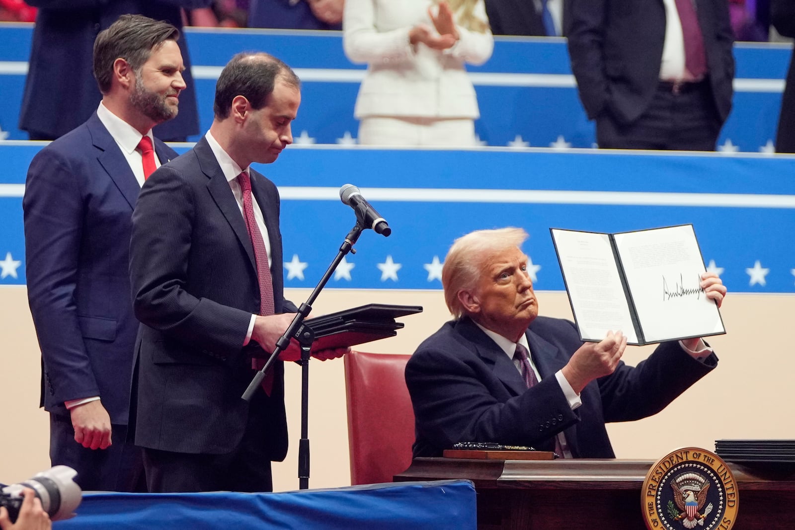 President Donald Trump signs executive orders on stage at an indoor Presidential Inauguration parade event in Washington, Monday, Jan. 20, 2025. (AP Photo/Mark Schiefelbein)