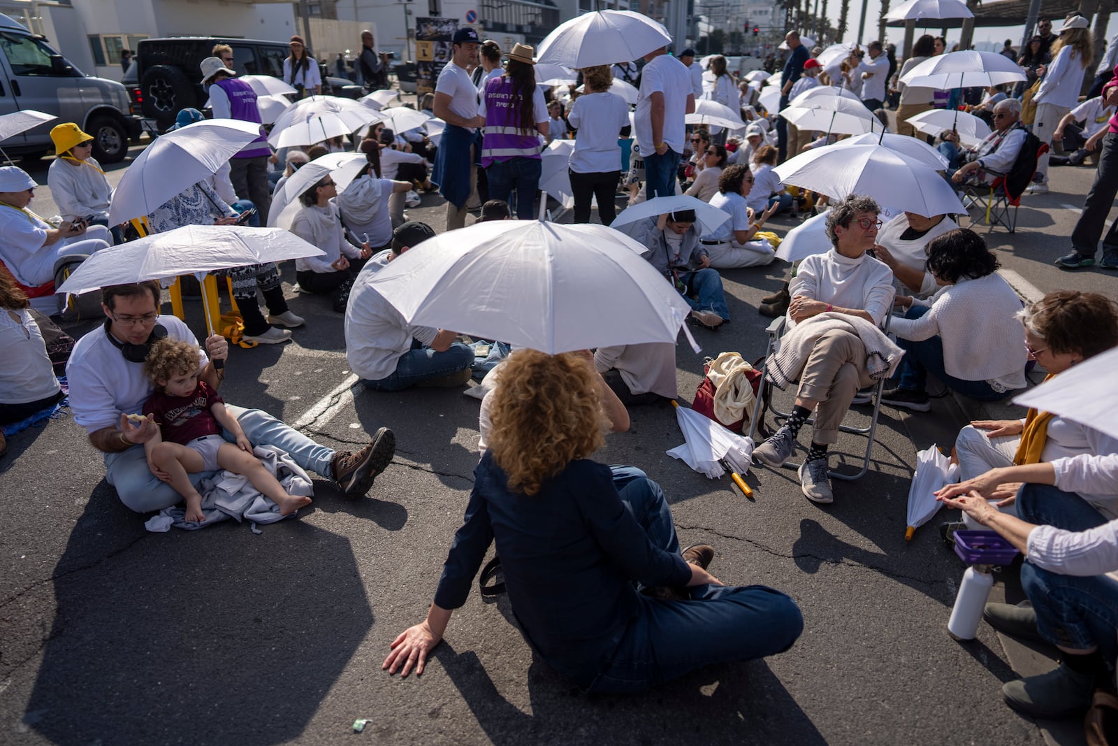 Activists sit on a road with white umbrellas during a protest calling for the release of hostages held in the Gaza Strip, in front of the U.S. Embassy branch office in Tel Aviv, Israel, Friday, Jan. 31, 2025. (AP Photo/Oded Balilty)