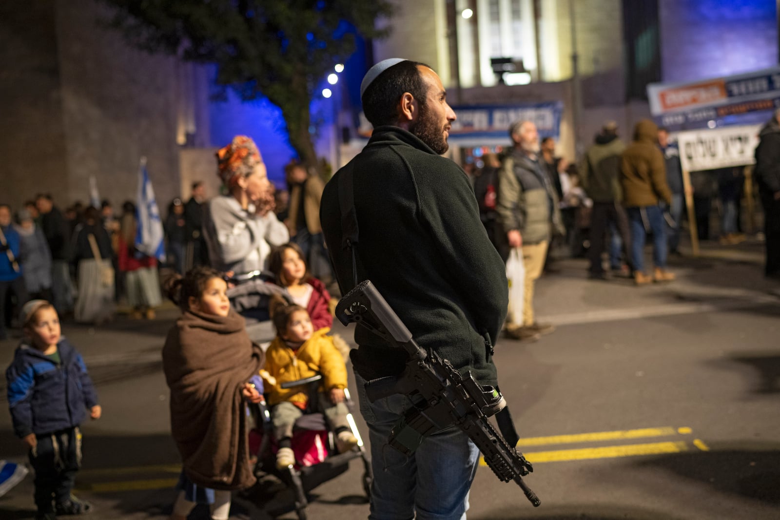 Carrying his weapon a man stands next to his family as he takes part in a protest demanding the continuation of the war against Hamas and the resettlement of the Gaza Strip, in Jerusalem, Thursday, Feb. 27, 2025. (AP Photo/Leo Correa)