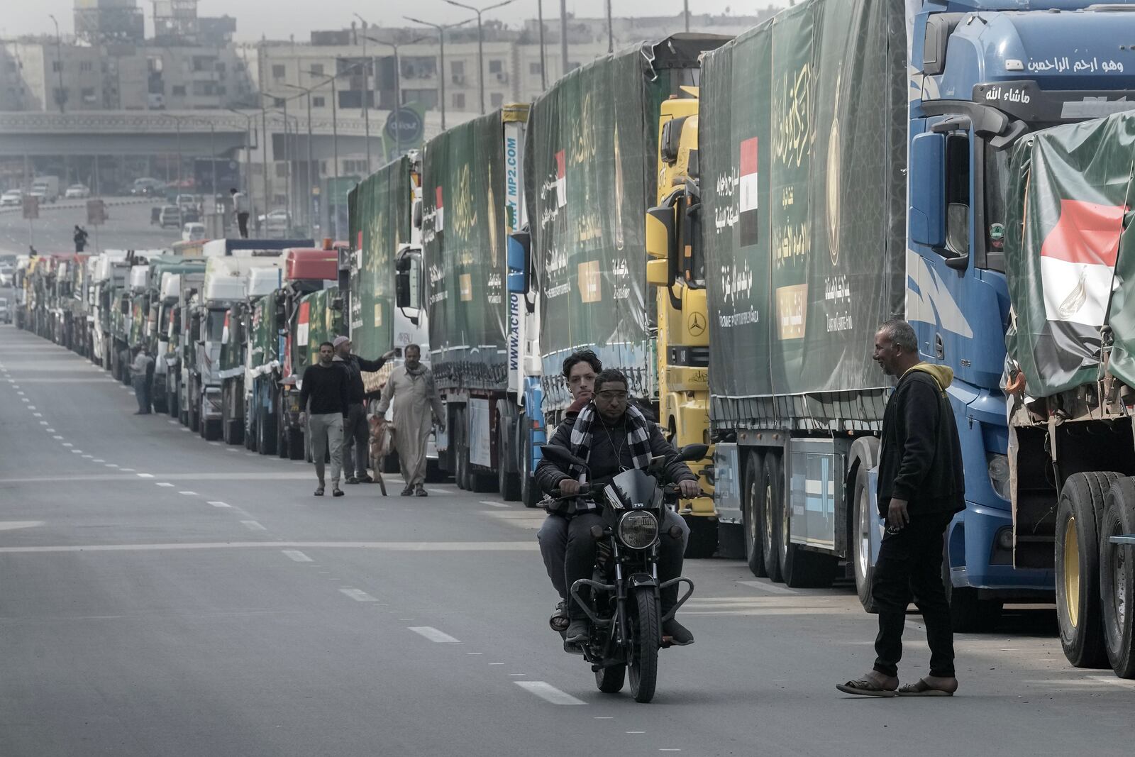 People walk past trucks of humanitarian aid at a parking point in Cairo, Egypt, as they wait to travel to cross the Rafah border crossing between Egypt and the Gaza Strip, Sunday, Jan. 26, 2025. (AP Photo/Amr Nabil)