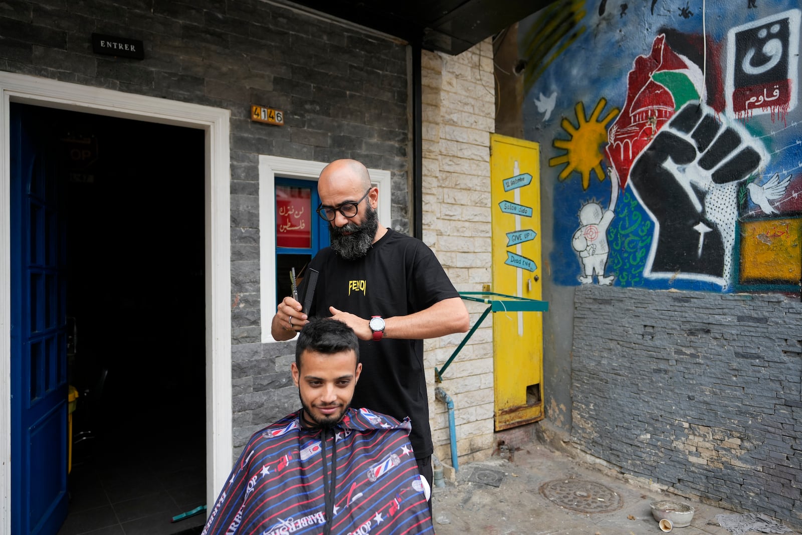 A Lebanese barber cuts the hair of a customer in front of his shop in Dahiyeh, Beirut, Lebanon, Wednesday, Oct. 16, 2024. The Arabic placard on the window reads: "We will not leave Palestine." (AP Photo/Hassan Ammar)