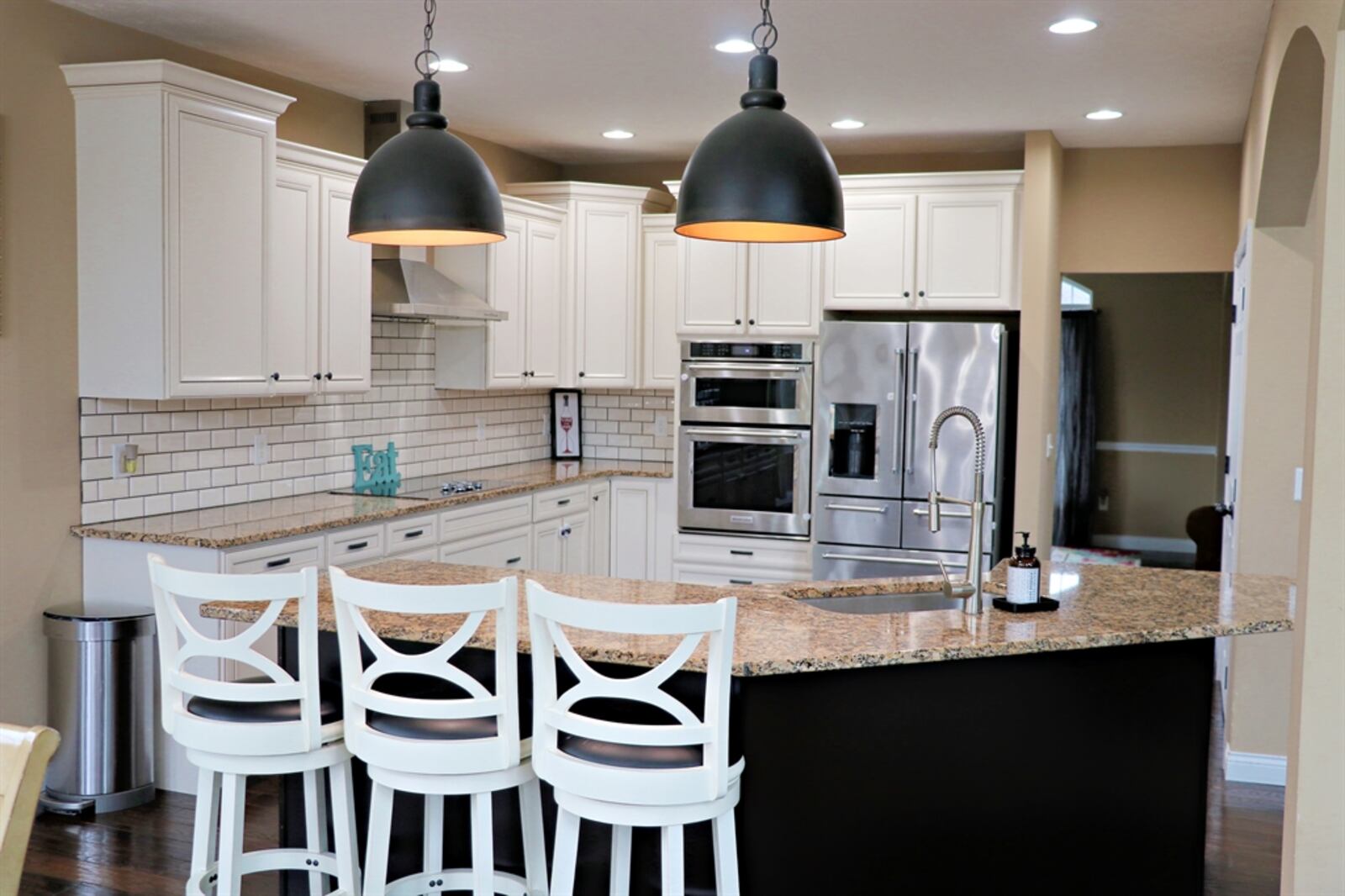 The L-shaped kitchen has antique-white cabinetry that wraps around stainless-steel appliances, including a glass cooktop and double-wall ovens. Subway tile complements the cabinetry with the granite counters. 