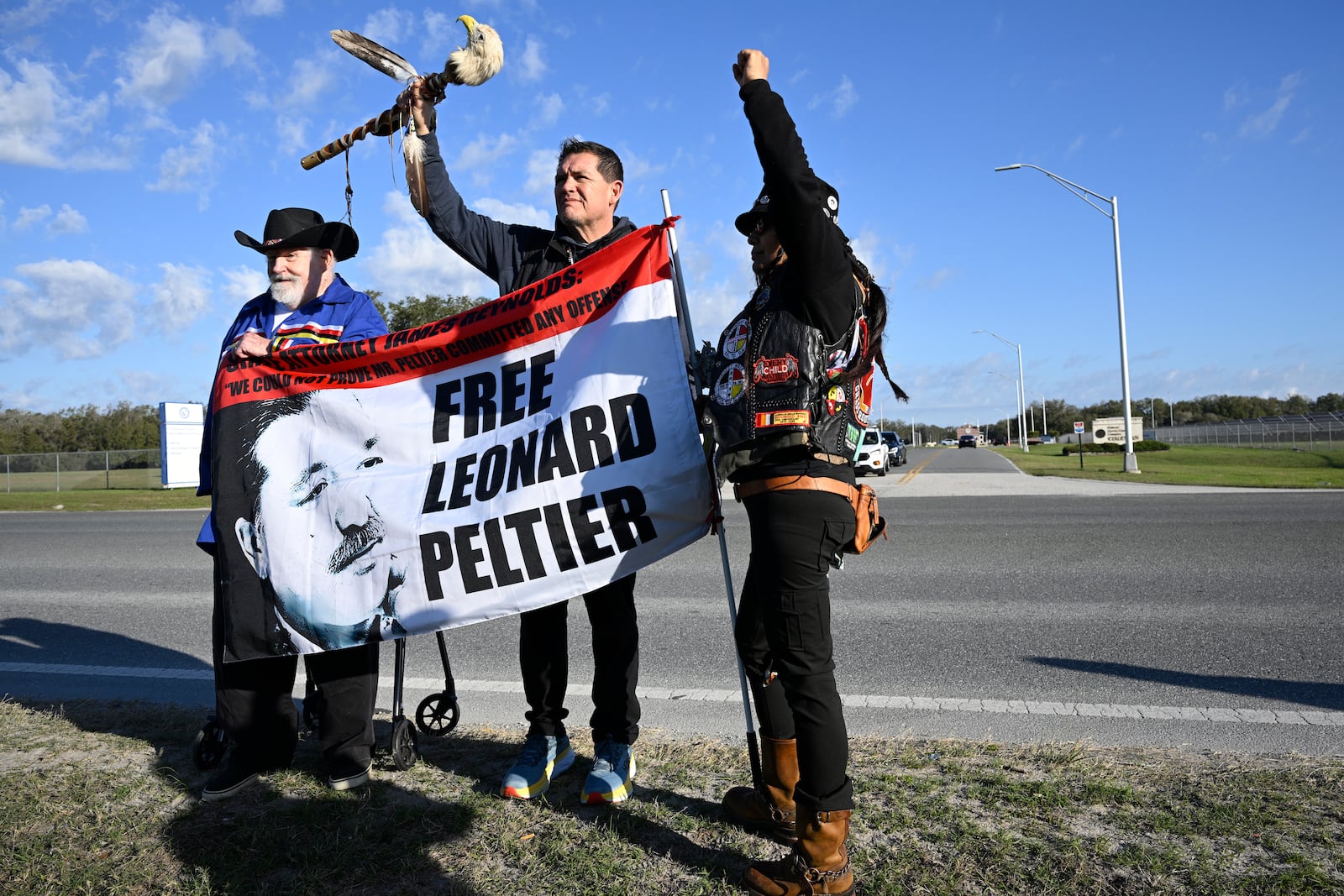 Supporters Mike McBride, left, Ray St. Clair, center, and Tracker Gina Marie Rangel Quinones stand in front of Federal Correctional Complex, Coleman, while awaiting the release Leonard Peltier, Tuesday, Feb. 18, 2025, in Sumterville, Fla. (AP Photo/Phelan M. Ebenhack)