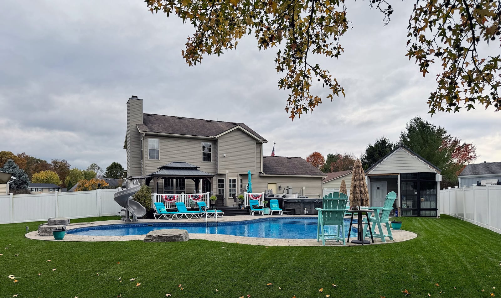 The rear has a vinyl privacy fence, a saltwater lagoon-style pool with slide, a pool house with bar and a wooden deck with railings connected to a stamped concrete patio and pool surround.