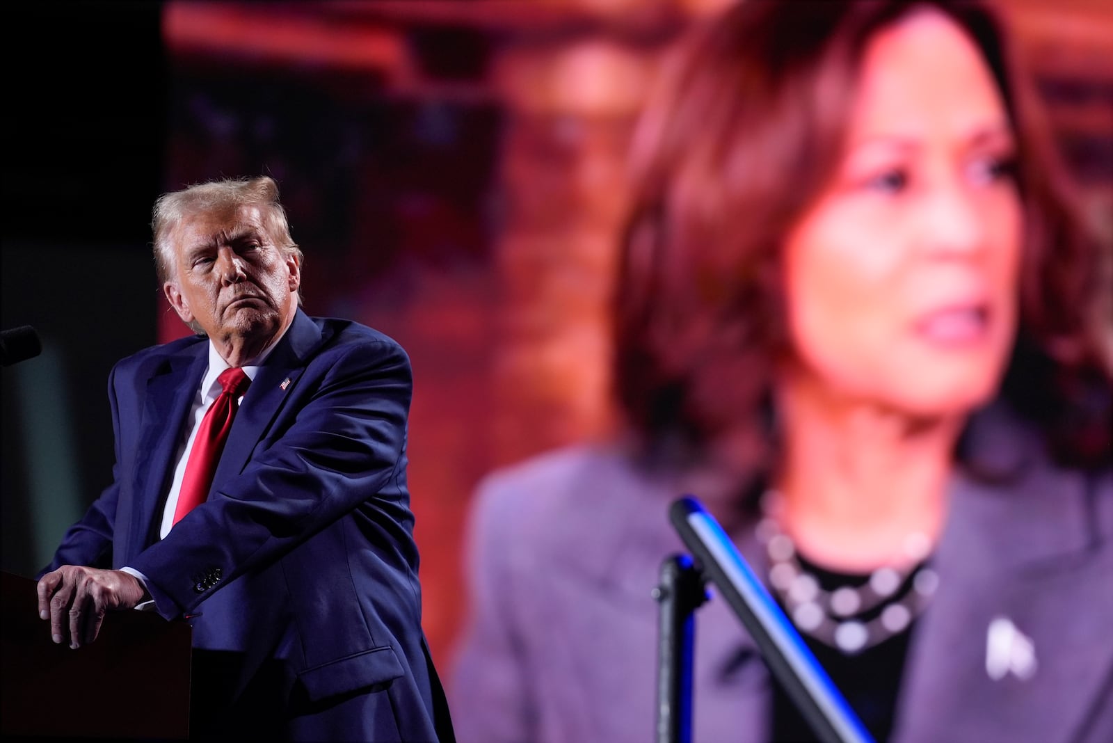 Republican presidential nominee former President Donald Trump watches a video on a screen with Democratic presidential nominee Vice President Kamala Harris as he speaks during a campaign event, Friday, Oct. 18, 2024 in Detroit. (AP Photo/Carlos Osorio)