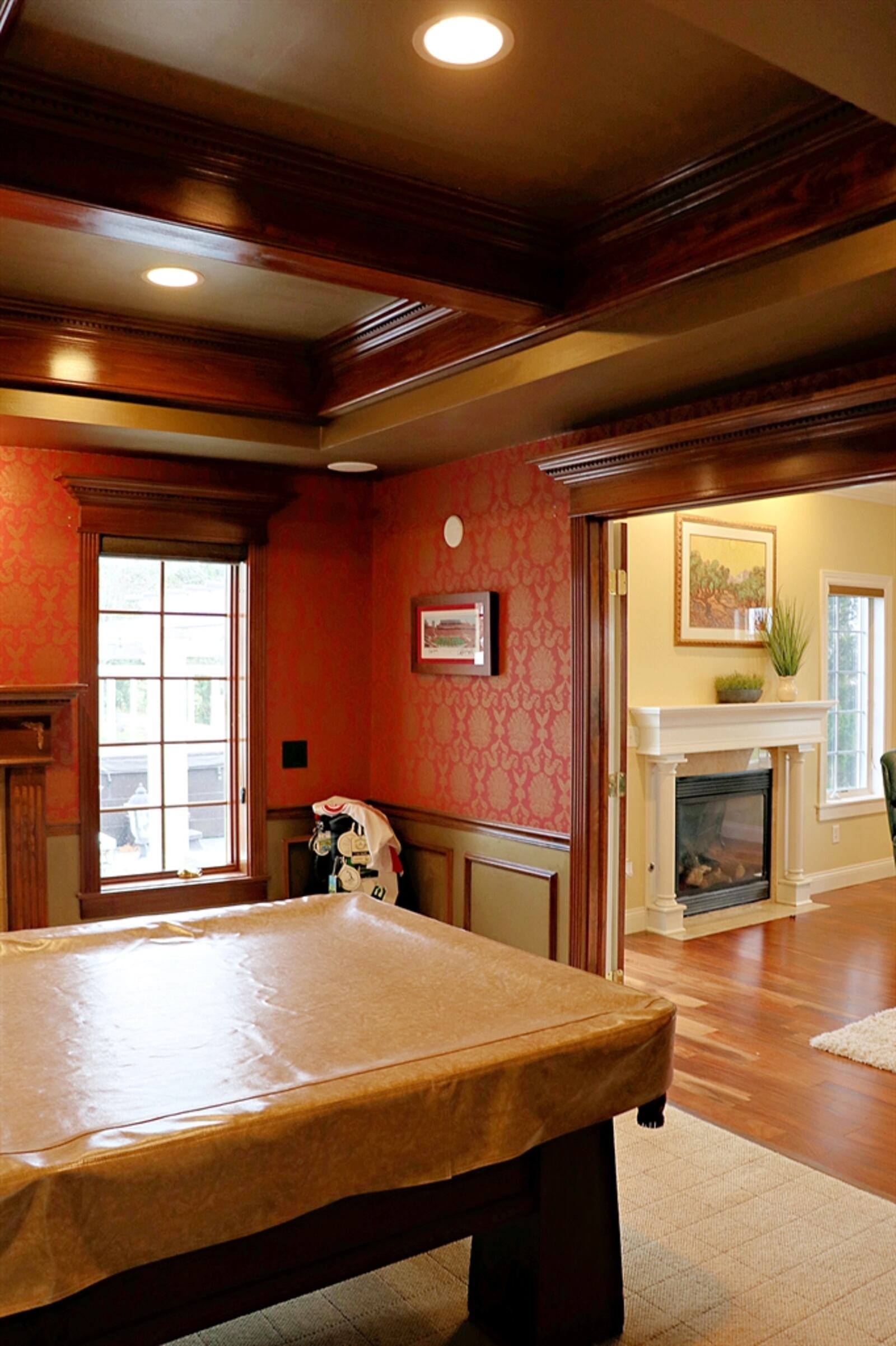 French doors open from the great room into a billiard room or library. A coffered ceiling has additional lighting and complements the wainscoting. Windows with wood trim flank the fireplace.