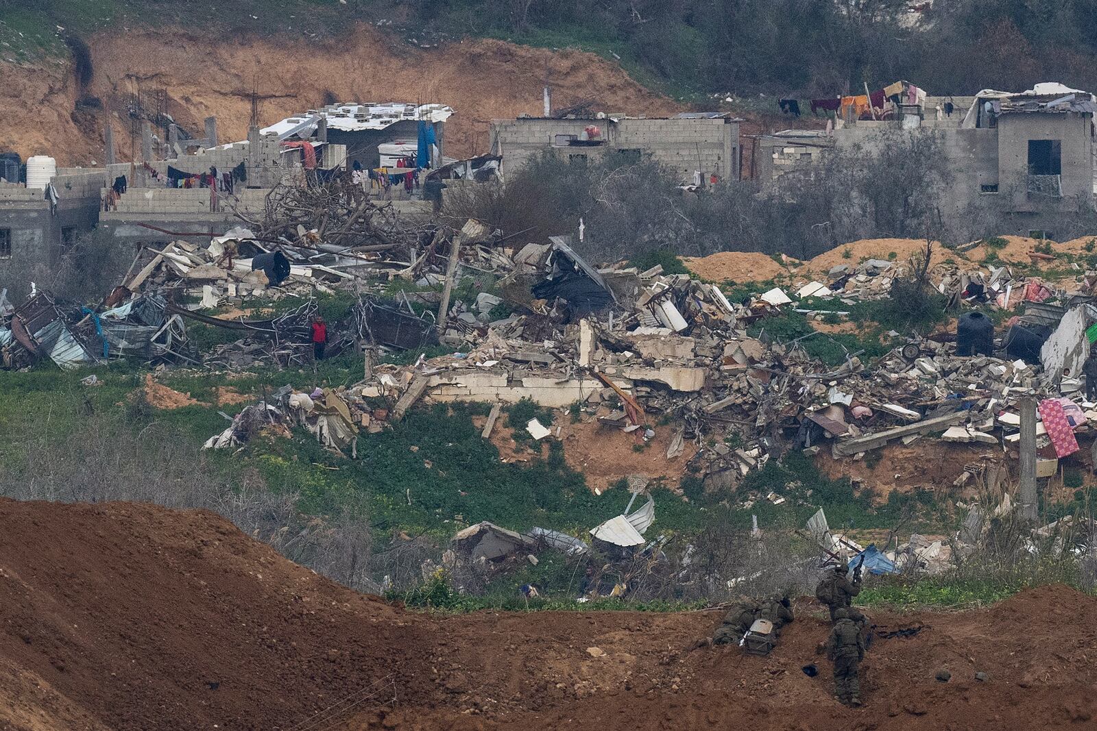 A Palestinian man wearing a red shirt, left, stands amidst the rubble of destroyed buildings, watching Israeli soldiers, bottom right, take position in the northern Gaza Strip, as seen from southern Israel, Sunday, Feb. 9, 2025. (AP Photo/Ohad Zwigenberg)