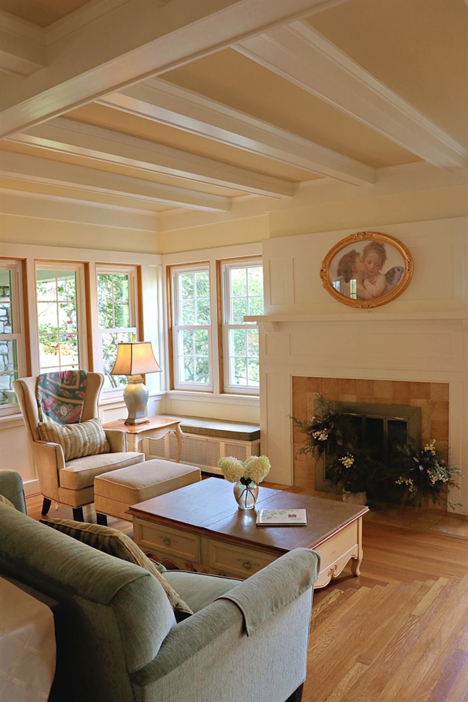 French doors open from the foyer into the living room, which has a coffered ceiling. The wood beams have been painted white to lighten the room and several windows fill the space with natural light. A fireplace has a stone-tile surround and hearth.