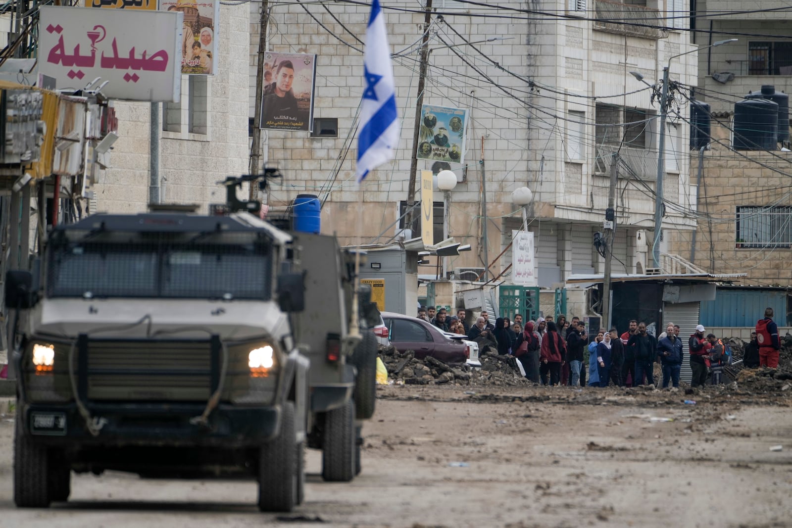 Palestinians look at Israeli military vehicles guard a road where a military bulldozer operates in the West Bank refugee camp of Jenin Wednesday, Jan. 22, 2025. (AP Photo/Majdi Mohammed)