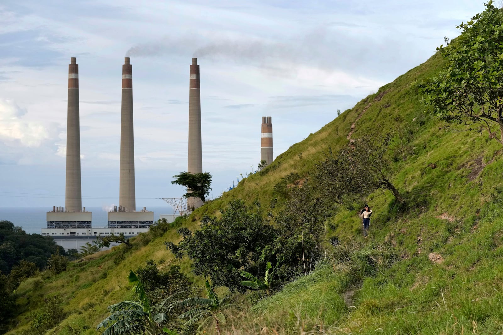 FILE - A couple walk on a hill called 'Teletubbies Hill', a locally popular tourist attraction, as the chimneys of Suralaya coal power plant looms in the background, in Cilegon, Indonesia, on Jan. 8, 2023. (AP Photo/Dita Alangkara, File)