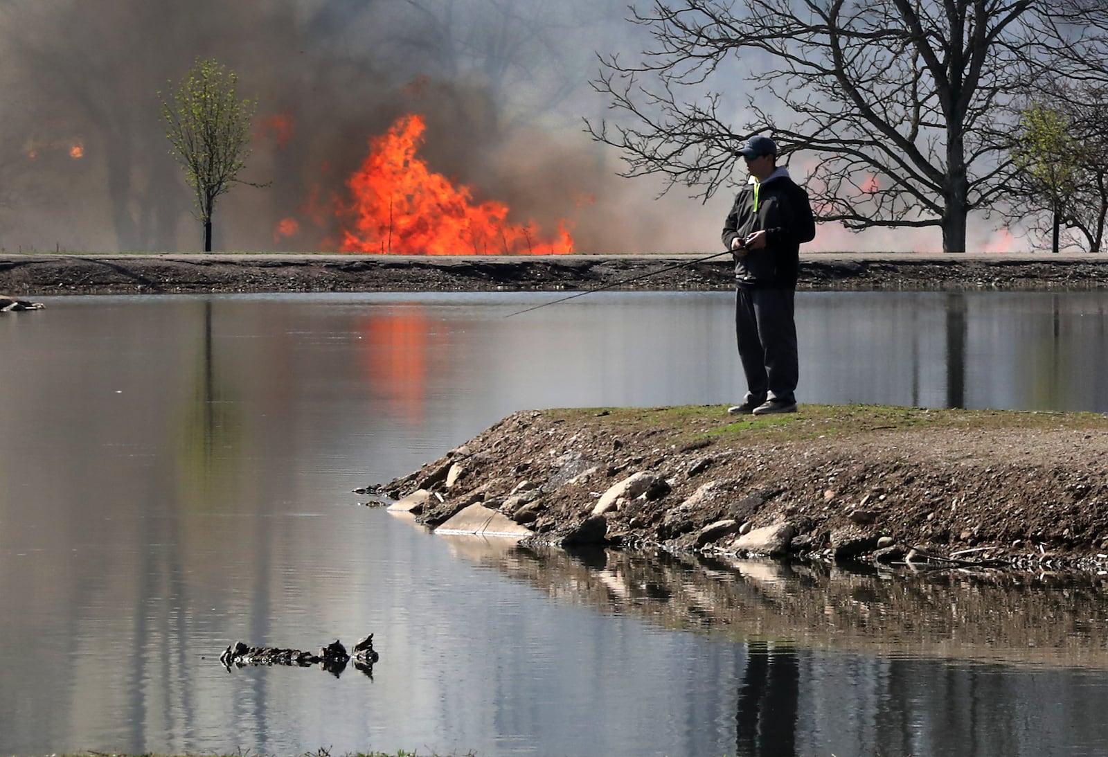 A fisherman takes advantage of the warm weather and sunshine Friday at Old Reid Park while a field fire rages across the lake. The fire was a controled burn by National Trail Parks and Recreatiion who were burning the tall grass in the Kirby Preserve. BILL LACKEY/STAFF