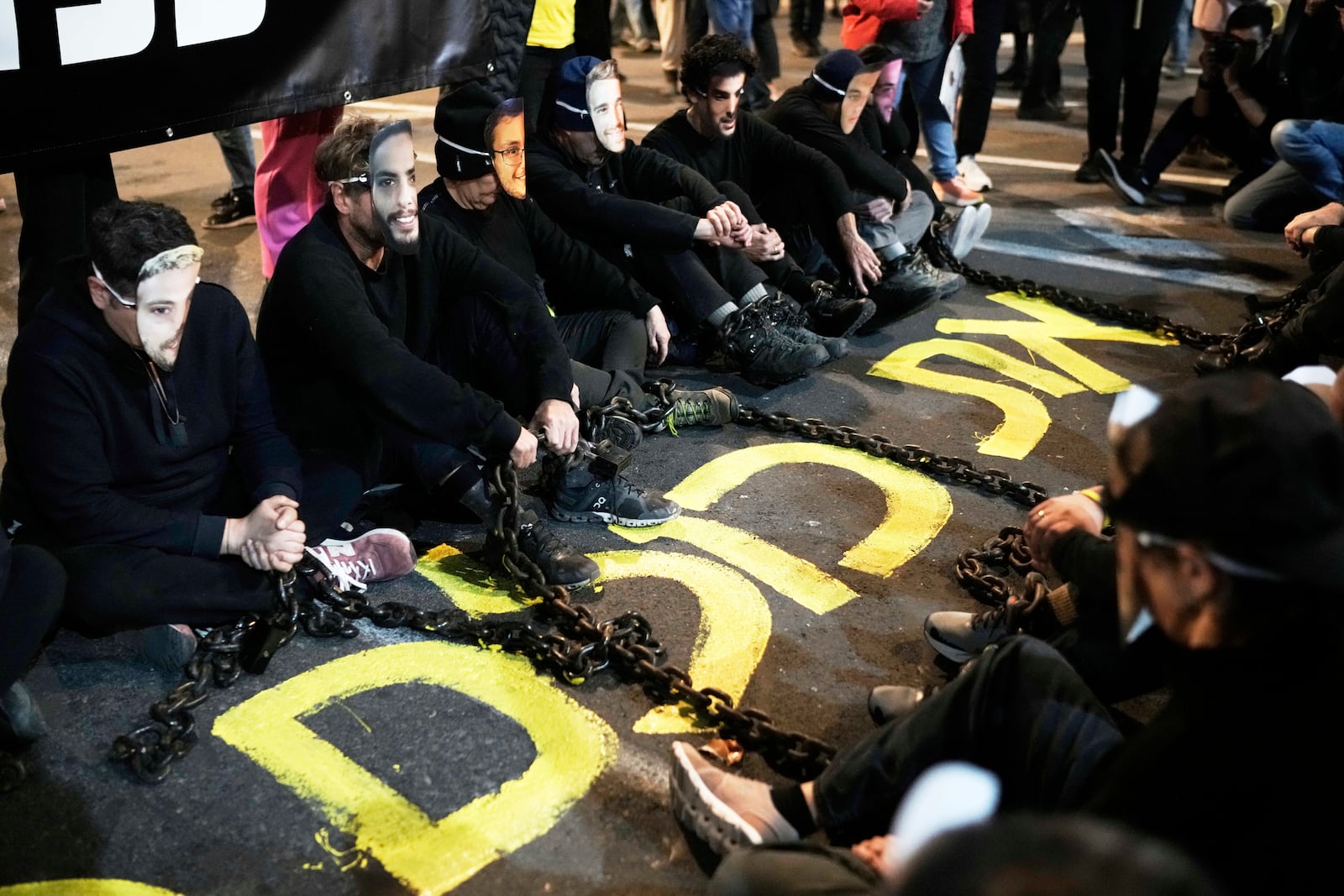 Activists sit on a street with iron chains as they wear masks depicting the faces of hostages held by Hamas in the Gaza Strip during a protest demanding their immediate release, in Tel Aviv, Israel, Saturday, March 1, 2025. Hebrew reads: "all of them." (AP Photo/Leo Correa)