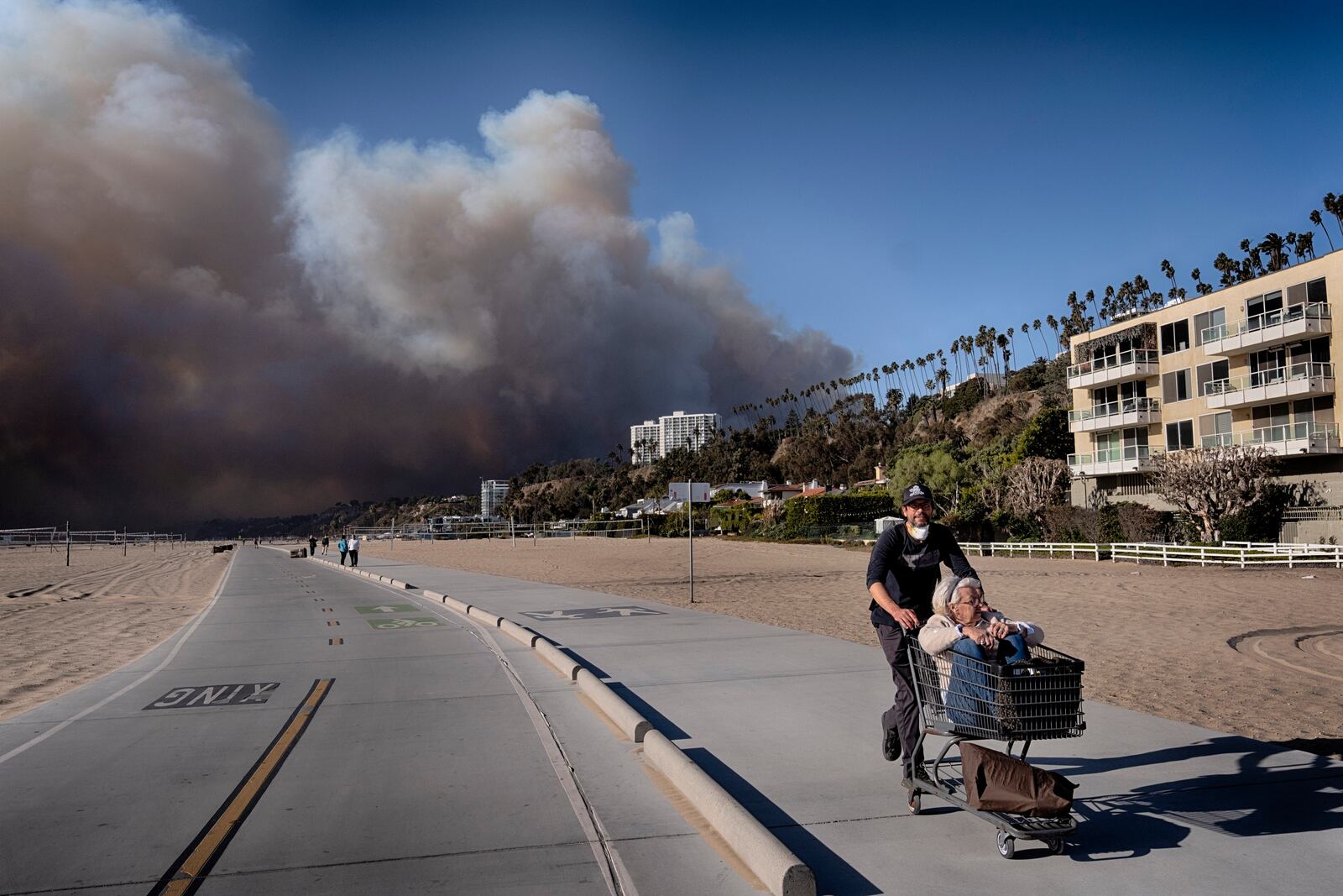 FILE - Jerome Krausse pushes his mother-in-law in a shopping cart as they evacuate from their home in the Pacific Palisades after a wildfire swept through their neighborhood in Santa Monica, Calif., on Tuesday, Jan. 7, 2025. (AP Photo/Richard Vogel, File)