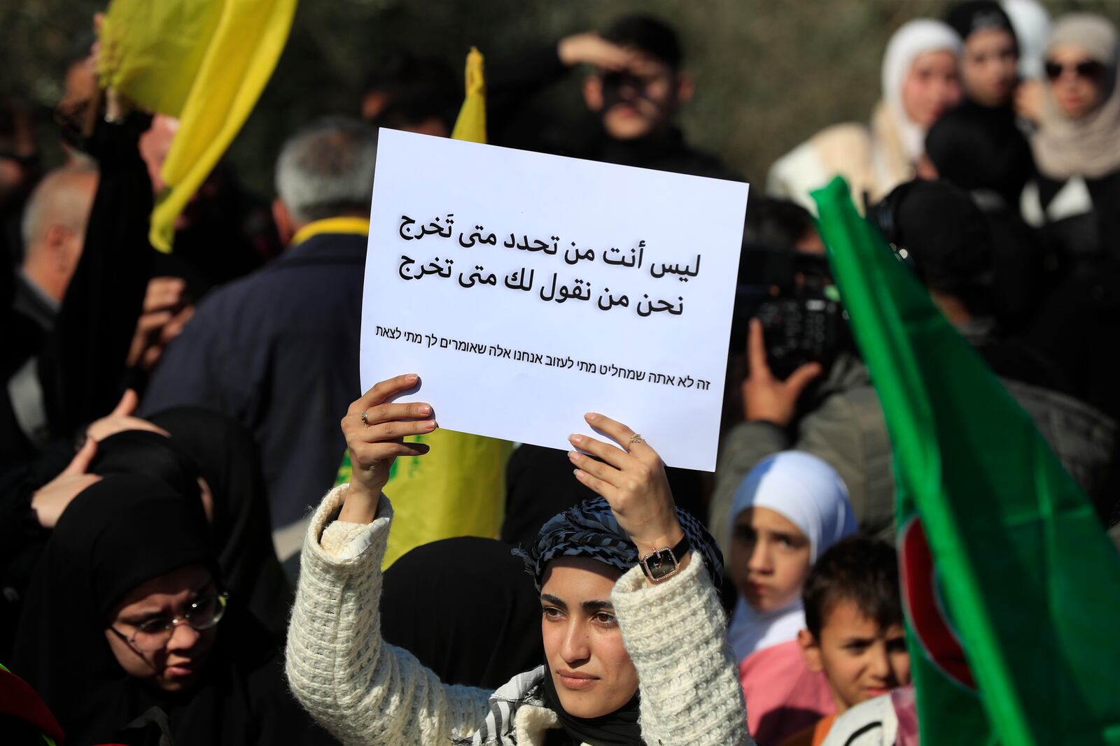 A Lebanese woman carries an Arabic placard that reads:"It's not you who decides when to go out, we tell you when to go out," as she gathers with other citizens who prepare to return to their villages, while Israeli soldiers block a road leading to the southern Lebanese village of Kfar Kila, Lebanon, Sunday, Feb. 2, 2025. (AP Photo/Mohammed Zaatari)