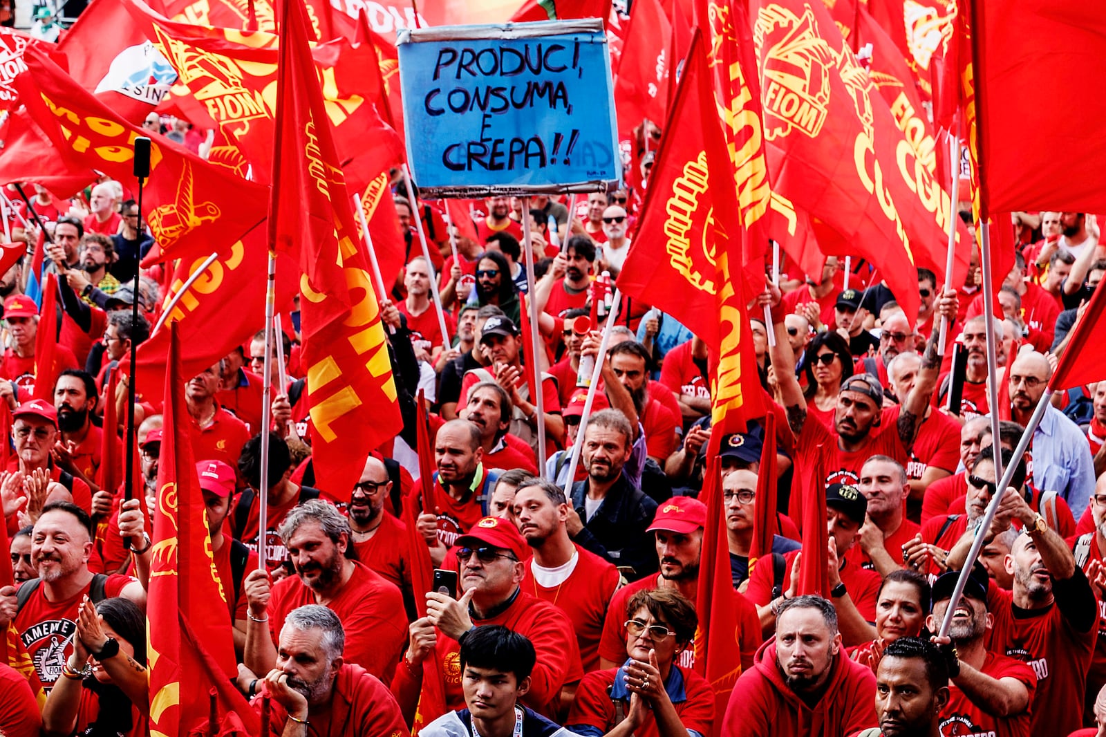 Demonstrators wave union flags on the occasion of a national strike of metalworkers of the automotive sector for the first time in 20 years, in Rome, Friday, Oct. 18 2024. Writing on sign at center reads in Italian "Produce, Consume and Die!". (Roberto Monaldo/LaPresse via AP)