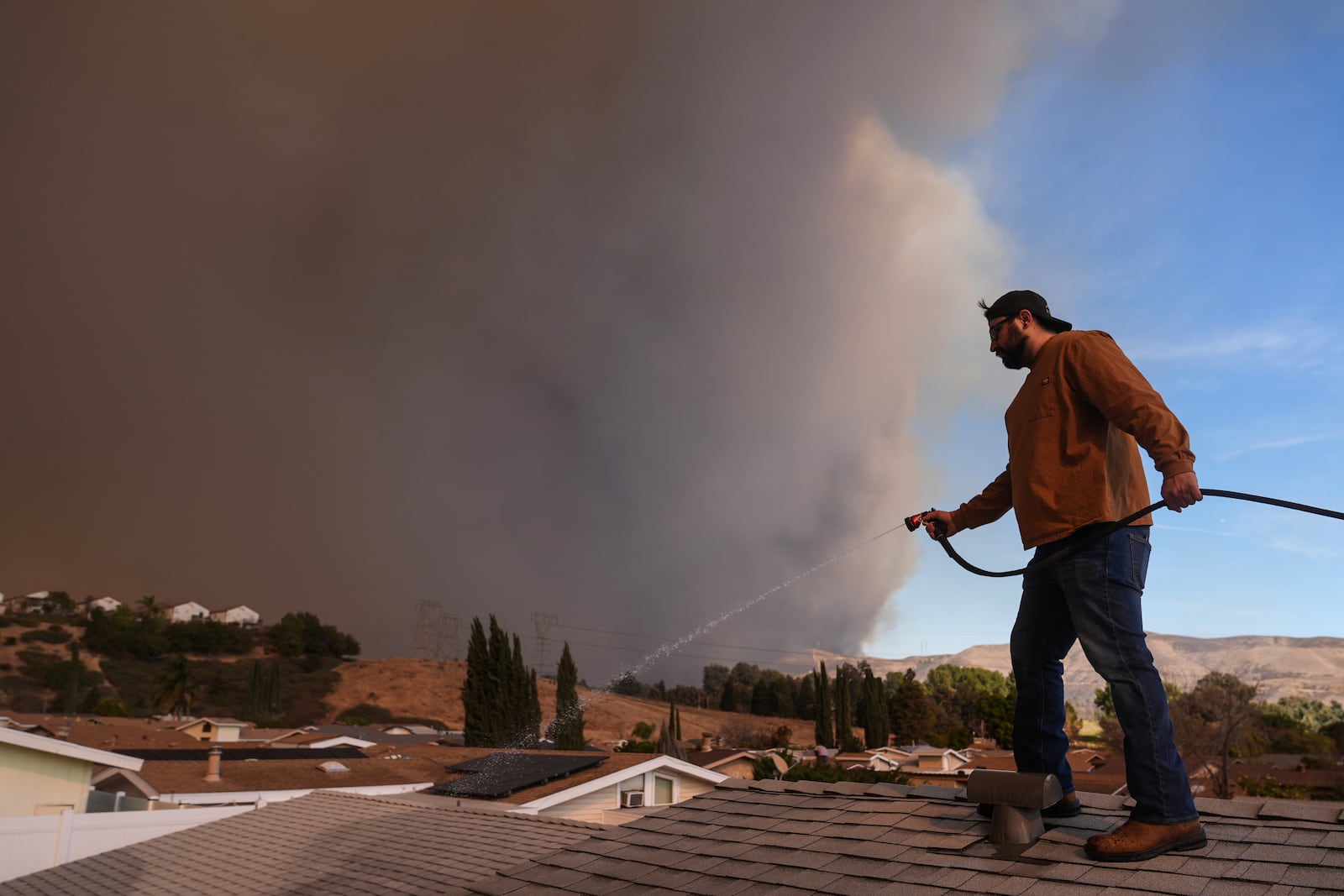 Andrew Aguilar spray water from the top of the roof at his brother's home Castaic, Calif., as a large plume of smoke caused by the Hughes Fire rises from Castaic Lake Wednesday, Jan. 22, 2025. (AP Photo/Marcio Jose Sanchez)