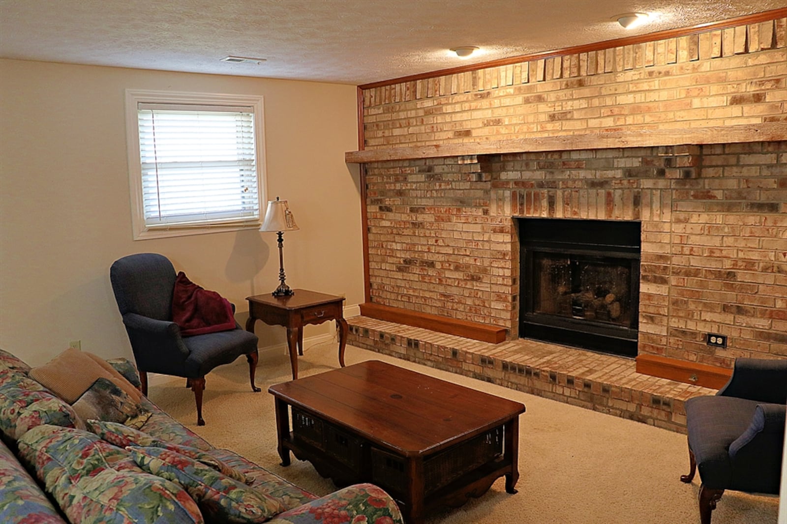 Two daylight windows fill the lower-level family room with natural light, and a brick gas fireplace stretches across one wall. The fireplace has a raised brick hearth and a wood-beam mantel.