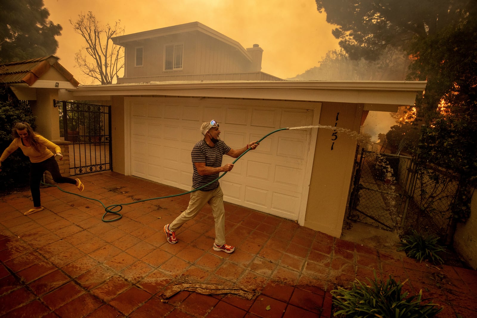 Will Adams uses a garden hose to keep flames from damaging his home as the Palisades Fire advances in the Pacific Palisades neighborhood of Los Angeles, Tuesday, Jan. 7, 2025. (AP Photo/Ethan Swope)